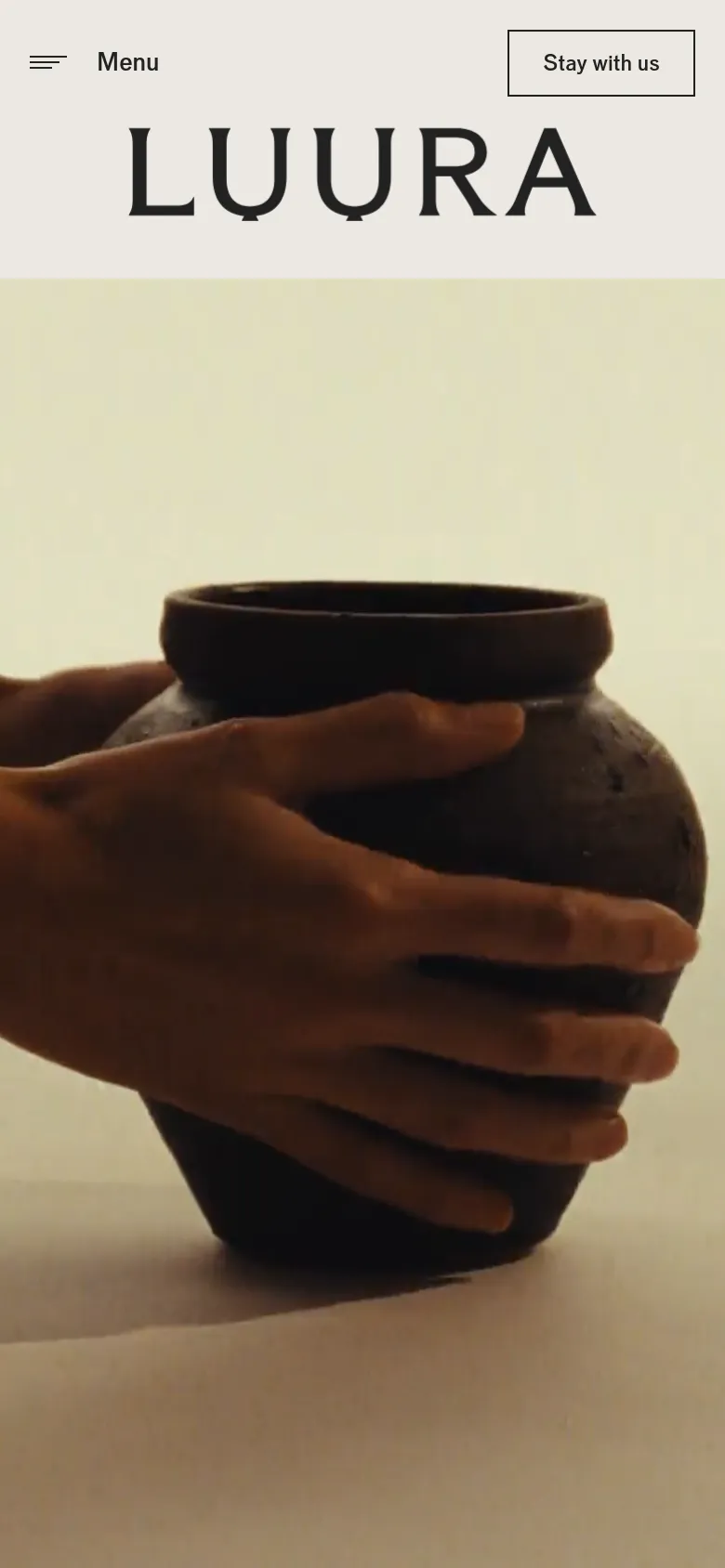 The mobile version maintains the minimalist cream aesthetic but reorganizes the header to include a hamburger menu icon labeled 'Menu' on the left and a 'Stay with us' call-to-action button on the right. The 'LUURA' logo is centered above a large, high-detail hero image of hands grasping a dark ceramic pot.