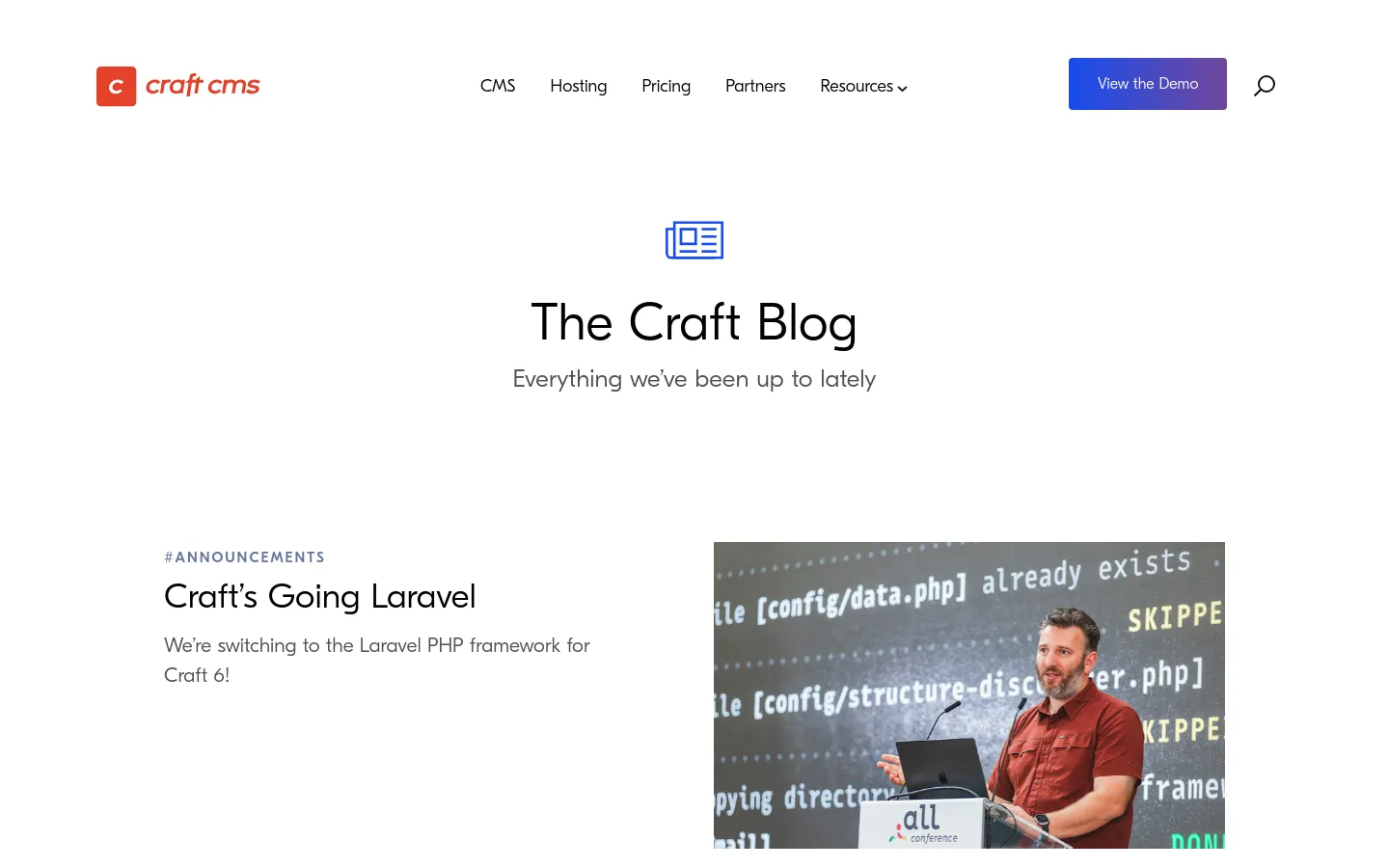 The Craft Blog landing page features a clean, white background with a centered header including a blue newspaper icon and the title 'The Craft Blog'. Below the header, a large featured article section displays a category tag '#ANNOUNCEMENTS', a headline 'Craft’s Going Laravel', a brief description, and a large photograph of a man speaking at a conference. A navigation bar at the top includes links for CMS, Hosting, Pricing, Partners, and Resources, alongside a 'View the Demo' button.