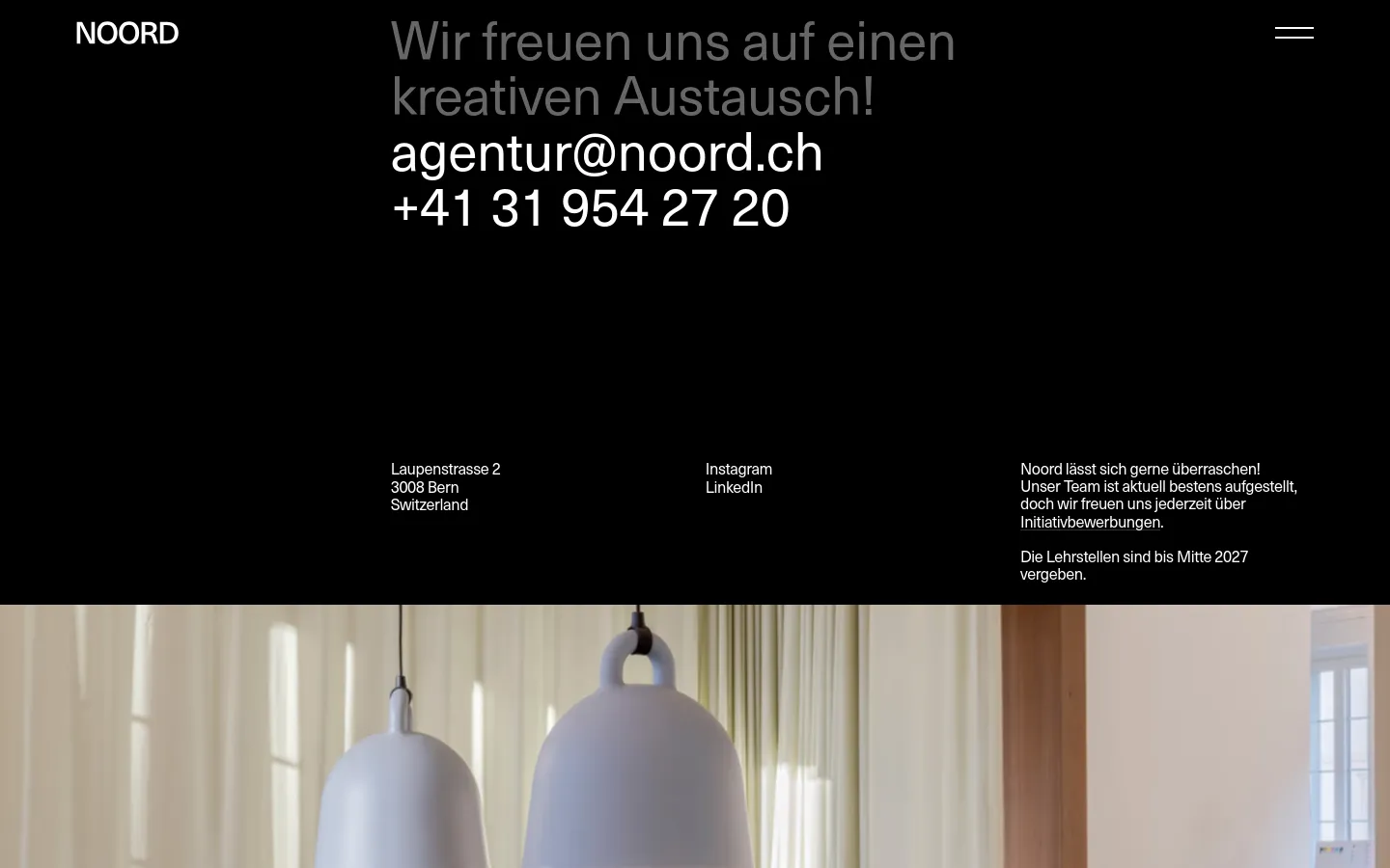 The desktop contact page features a minimalist black background with large white typography. A headline reads 'Wir freuen uns auf einen kreativen Austausch!' followed by the agency's email and phone number in a prominent font size. Below, contact details, social media links, and a brief note about job applications are arranged in three columns above a lifestyle photograph of hanging lamps.