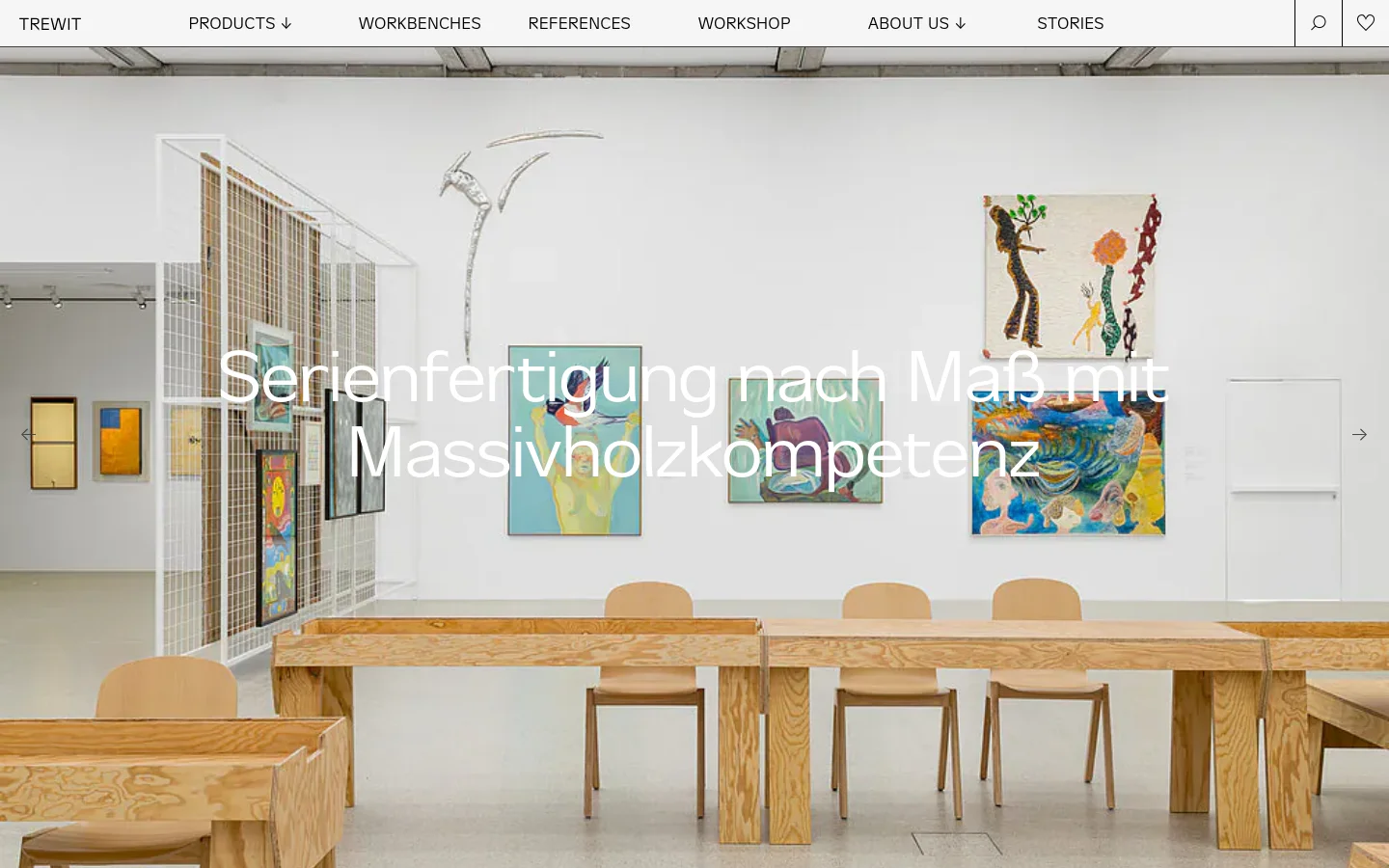 The desktop homepage features a minimalist design with a large hero image of a bright gallery space containing wooden tables, chairs, and colorful abstract paintings. A large white headline reads 'Serienfertigung nach Maß mit Massivholzkompetenz' centered over the image. A clean navigation bar at the top includes links for Products, Workbenches, References, Workshop, About Us, and Stories, alongside a search icon and a heart icon.