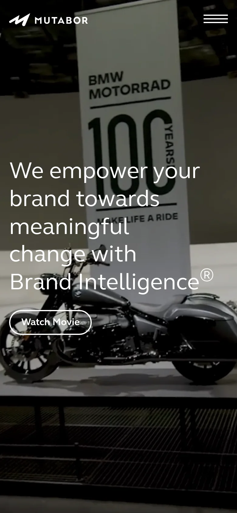 The mobile version uses a photographic background showing a BMW Motorrad display with a large '100' graphic. The same white headline and 'Watch Movie' button are overlaid on the image, with a hamburger menu icon in the top right corner.