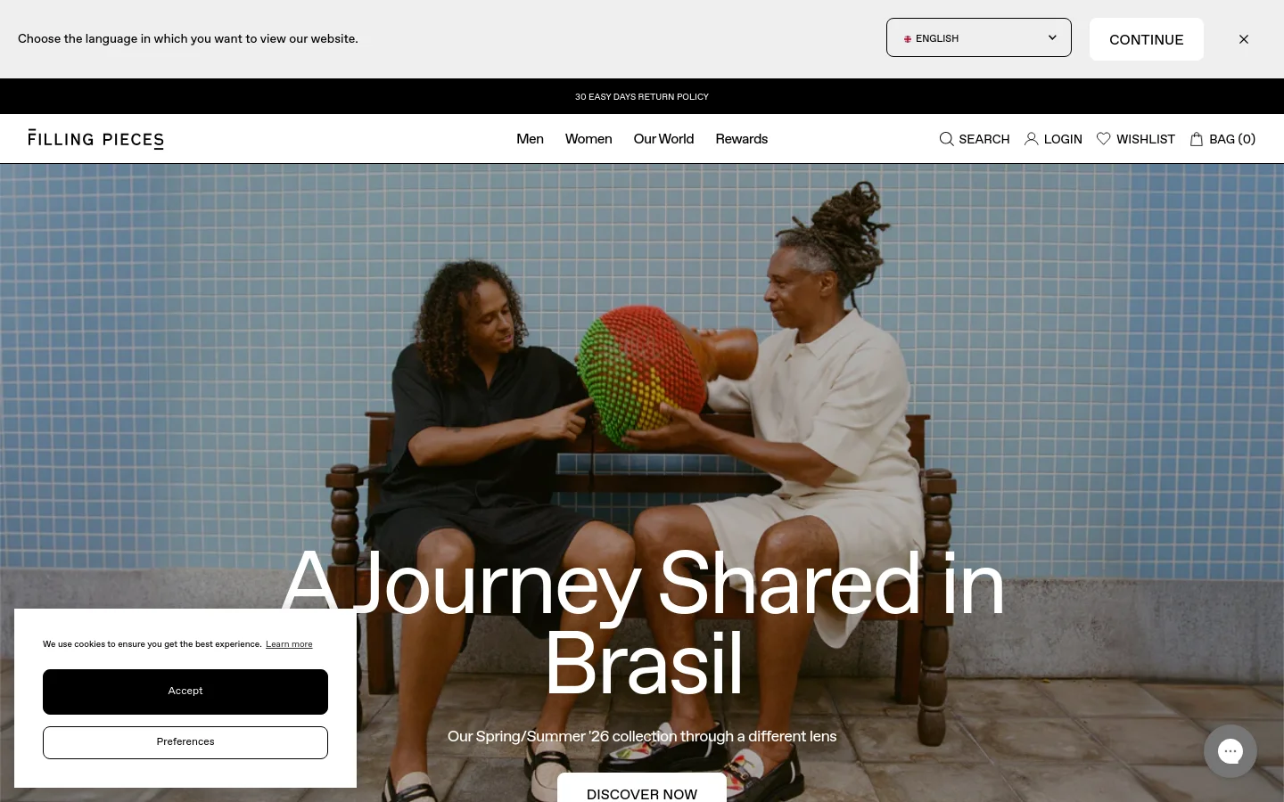 The desktop homepage features a large hero image of two people sitting on a bench against a blue tiled wall, overlaid with the headline 'A Journey Shared in Brasil' and a 'DISCOVER NOW' button. A top navigation bar includes links for Men, Women, Our World, and Rewards, alongside search, login, wishlist, and bag icons. A cookie consent banner and a language selection bar are visible at the top of the viewport.