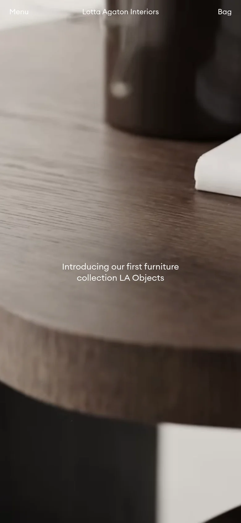 The mobile version uses the same dark wood grain hero imagery but simplifies the layout for a smaller screen. The navigation is condensed into a 'Menu' link on the left, the brand name remains centered, and 'Bag' is on the right. The central headline 'Introducing our first furniture collection LA Objects' is stacked vertically for better readability.