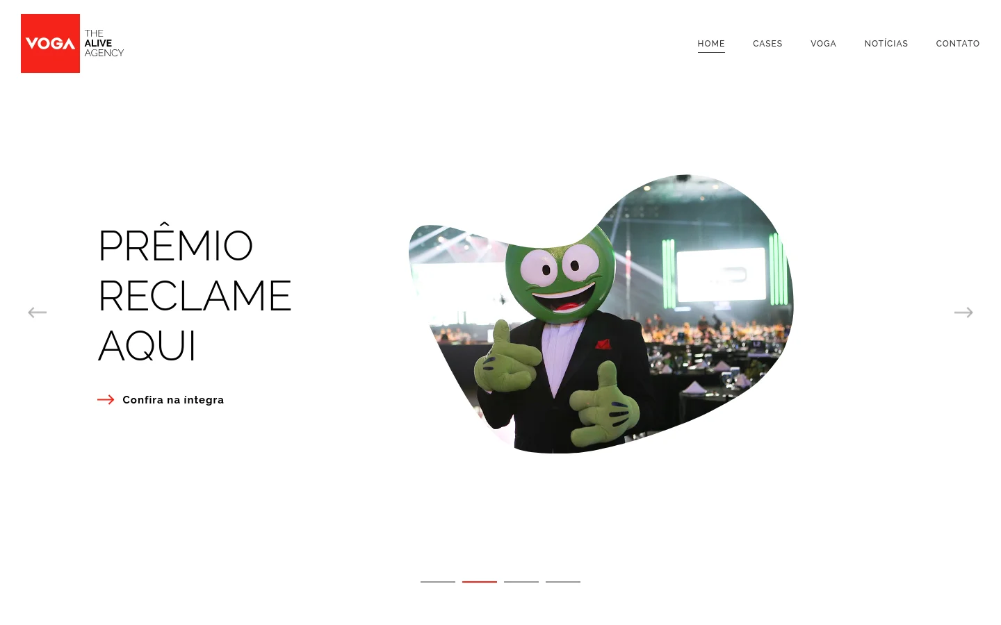 The desktop website for Voga agency features a minimalist white background with a clean navigation menu at the top right. A large, organic-shaped cutout displays a photo of a person in a green mascot suit, positioned next to the headline 'PRÊMIO RECLAME AQUI' and a red arrow call to action. A carousel indicator at the bottom shows the current slide position.