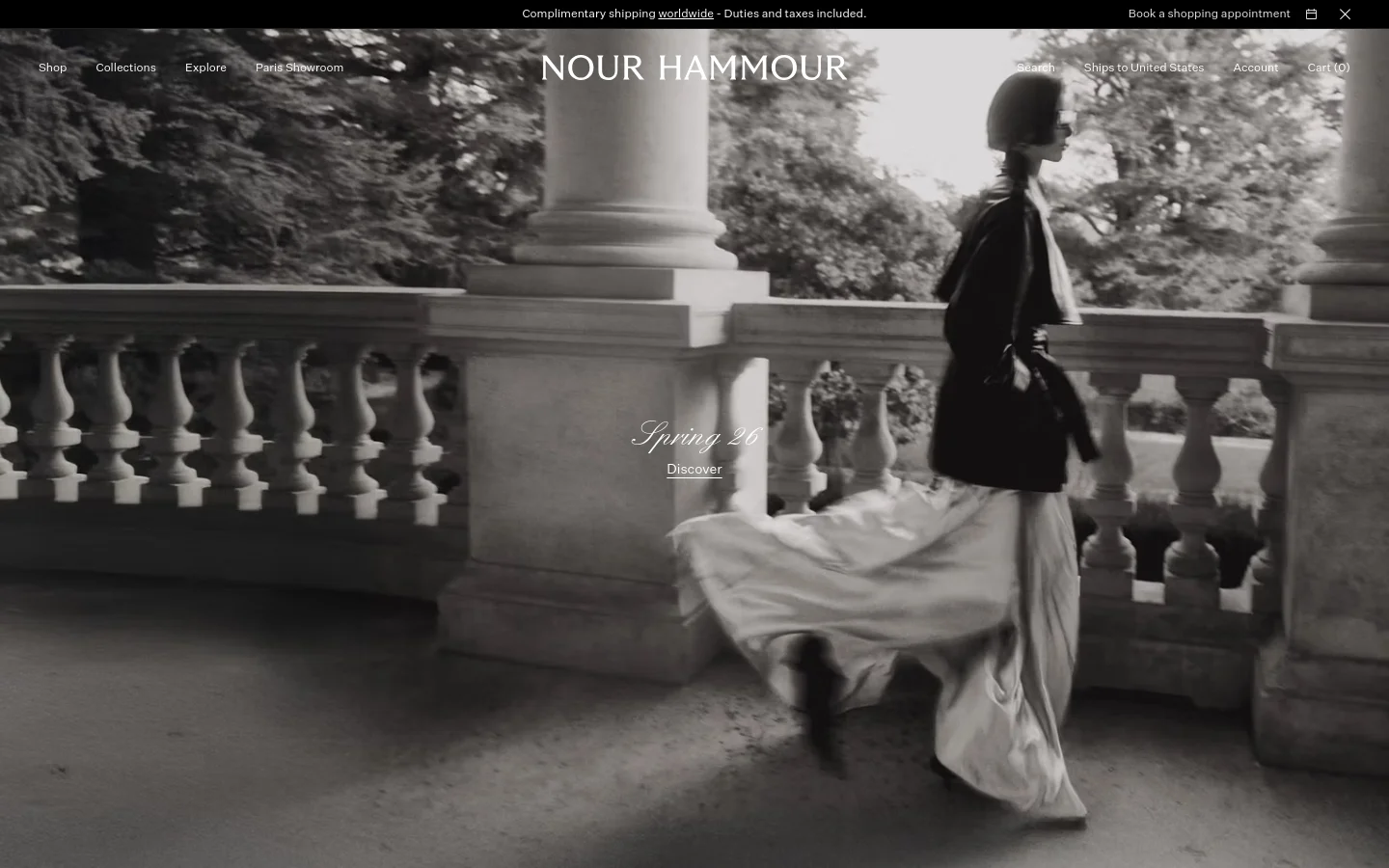 The desktop homepage features a large, black-and-white hero image of a person in flowing garments walking past a stone balustrade. The brand name 'NOUR HAMMOUR' is centered at the top, with a navigation menu including Shop, Collections, Explore, Paris Showroom, Search, Ships to United States, Account, and Cart (0). A central text overlay reads 'Spring 26' with a 'Discover' call to action.