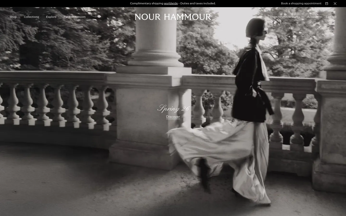 The desktop homepage features a large, black-and-white hero image of a person in flowing garments walking past a stone balustrade. The brand name 'NOUR HAMMOUR' is centered at the top, with a navigation menu including Shop, Collections, Explore, Paris Showroom, Search, Ships to United States, Account, and Cart (0). A central text overlay reads 'Spring 26' with a 'Discover' call to action.