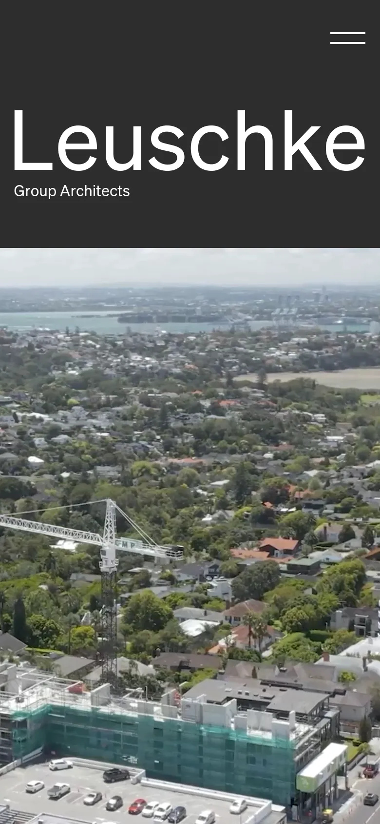 The mobile version uses a stacked layout where the large white 'Leuschke Group Architects' title sits atop a full-width aerial photograph of a coastal cityscape. The dark grey background from the desktop version is reduced to a header area containing a hamburger menu icon.