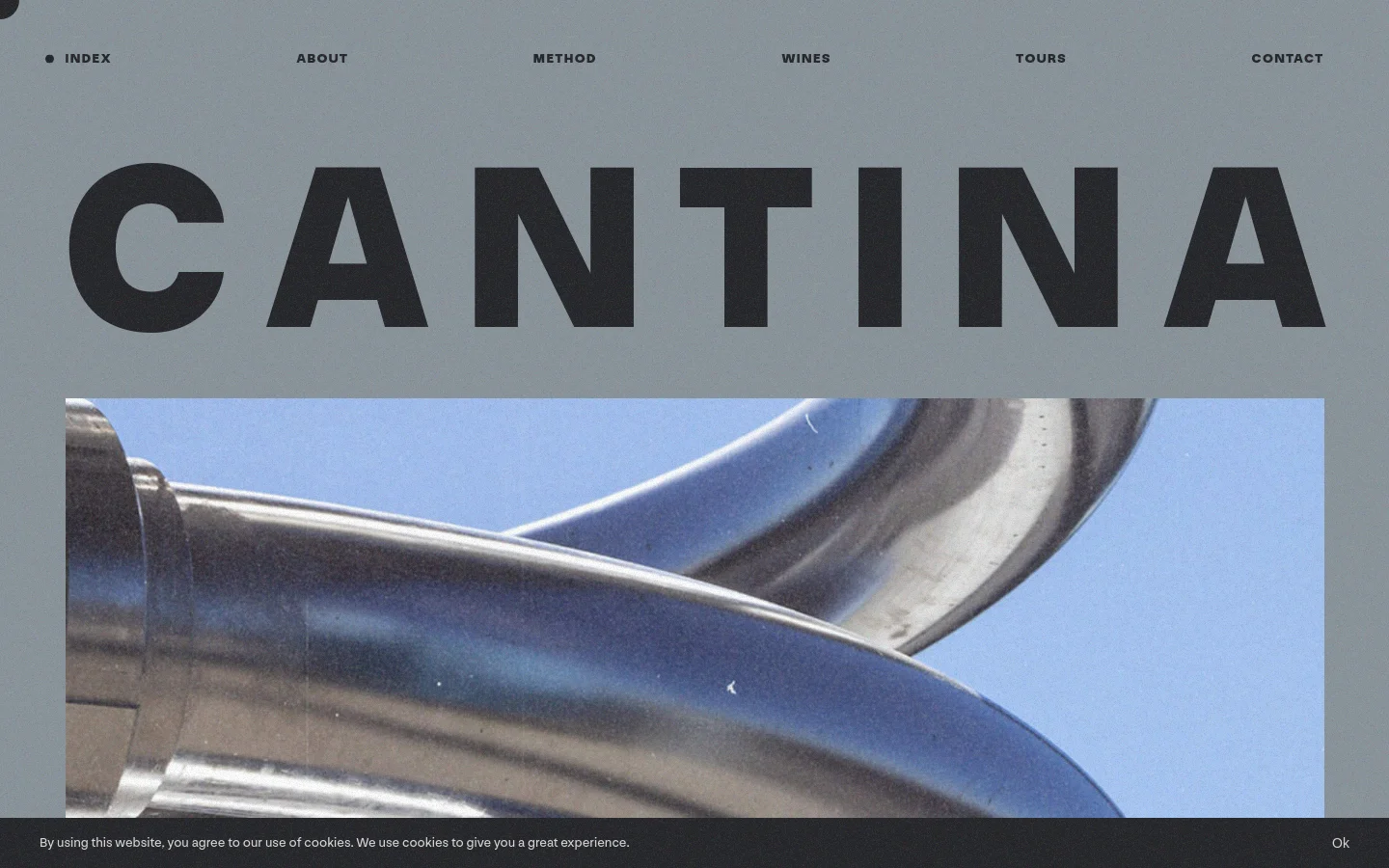 The desktop homepage features a minimalist gray background with a large, bold black headline reading 'CANTINA' above a wide cinematic image of polished stainless steel wine production equipment. A navigation menu at the top includes links for Index, About, Method, Wines, Tours, and Contact, while a cookie consent banner sits at the bottom.