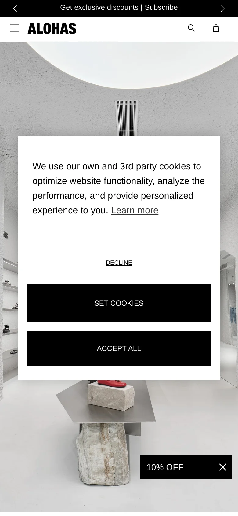 The mobile version presents a vertically oriented view of the white, textured boutique interior. The cookie consent overlay is prominent in the center of the screen, featuring large, stacked black buttons for 'SET COOKIES' and 'ACCEPT ALL'. A black promotional banner at the bottom reads '10% OFF' with a close icon, and the header includes a hamburger menu, logo, search icon, and shopping bag.