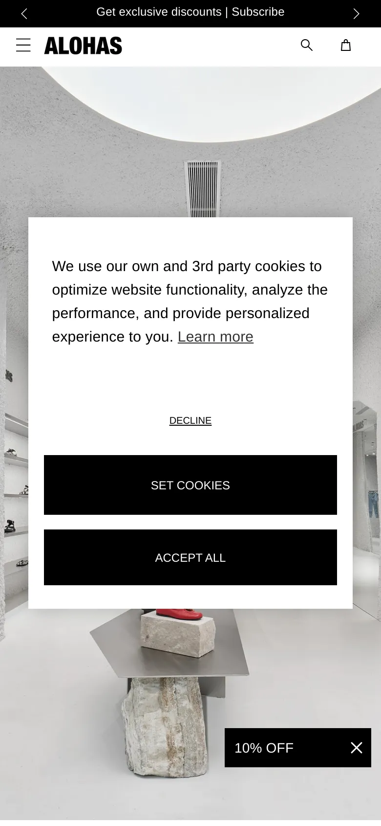 The mobile version presents a vertically oriented view of the white, textured boutique interior. The cookie consent overlay is prominent in the center of the screen, featuring large, stacked black buttons for 'SET COOKIES' and 'ACCEPT ALL'. A black promotional banner at the bottom reads '10% OFF' with a close icon, and the header includes a hamburger menu, logo, search icon, and shopping bag.