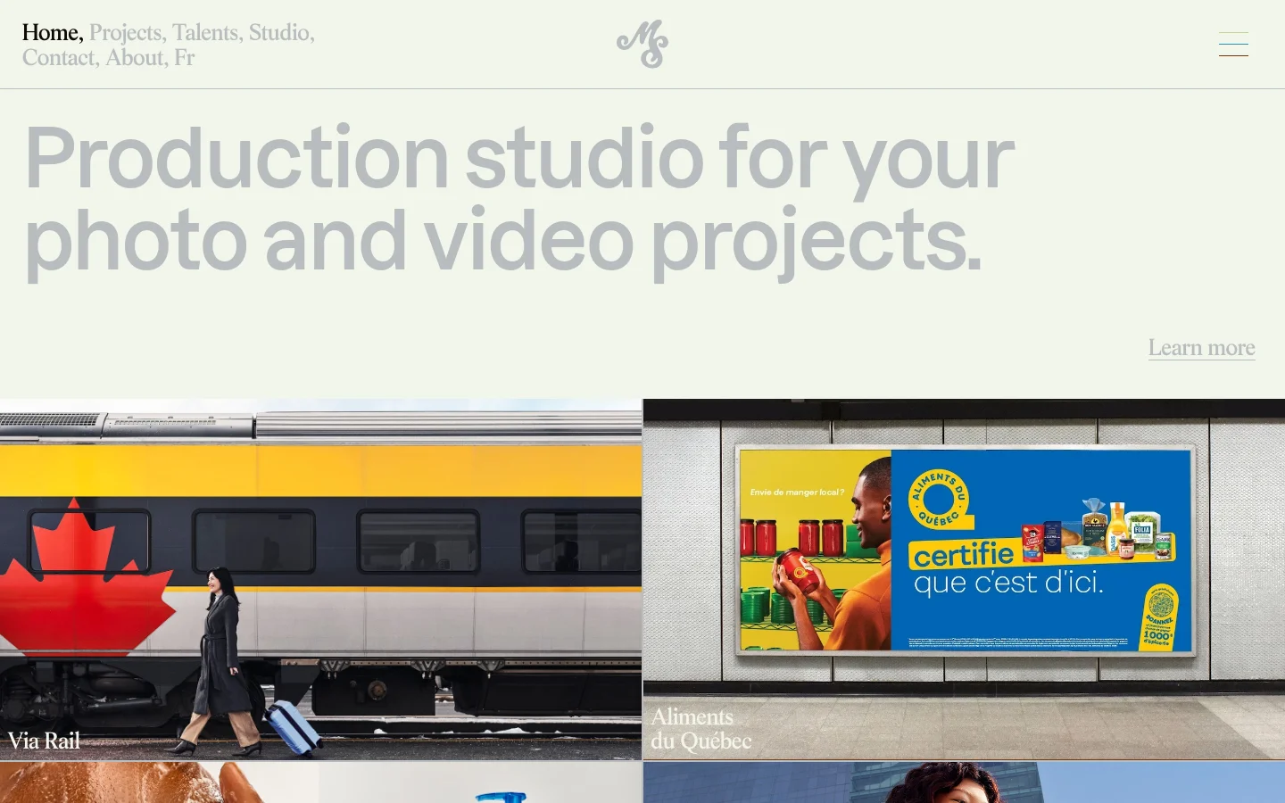 The desktop homepage features a minimalist design with a light beige background and large, bold grey typography stating 'Production studio for your photo and video projects.' A navigation menu is at the top left, a logo is centered, and a 'Learn more' link sits above a large split-screen hero image showing a person walking by a Via Rail train and a billboard for Aliments du Québec. The layout is spacious and clean.