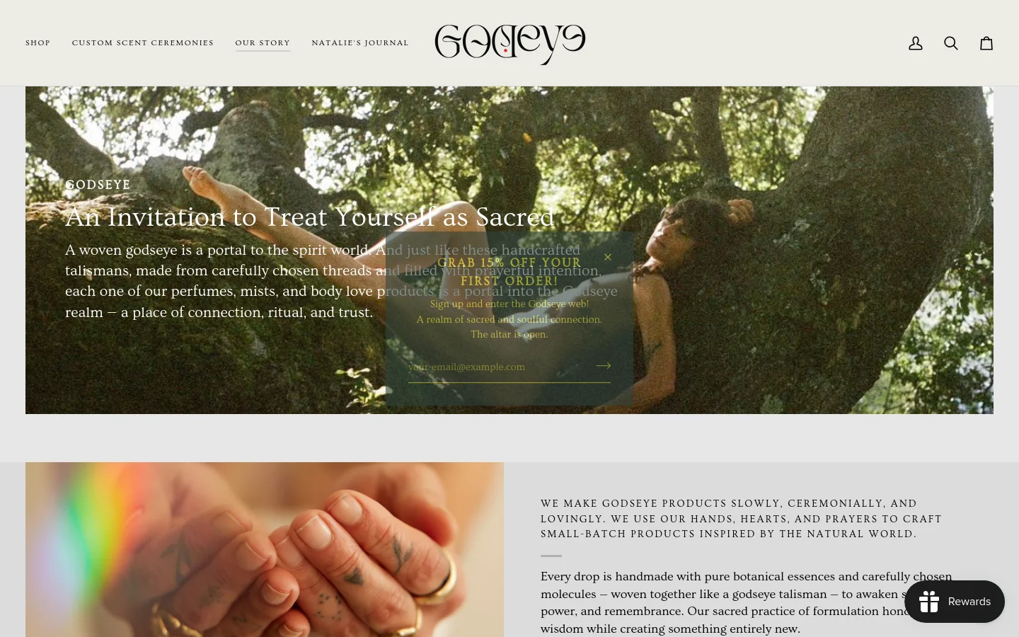 The desktop page features a large hero image of a person lying amidst lush green trees with white serif text overlay reading 'GODSEYE: An Invitation to Treat Yourself as Sacred.' Below the hero, a secondary section shows a close-up of hands with tattoos, paired with text describing the brand's slow, ceremonial creation process. The layout is airy with a clean white background and a minimalist navigation menu at the top.