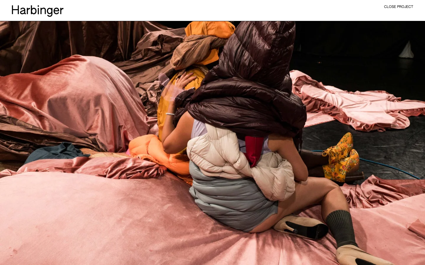 The Harbinger project page features a large, full-width hero image of a performer in voluminous, quilted clothing amidst pink and brown fabric. The headline 'Harbinger' is positioned at the top left, with a 'CLOSE PROJECT' link at the top right on a clean white background.