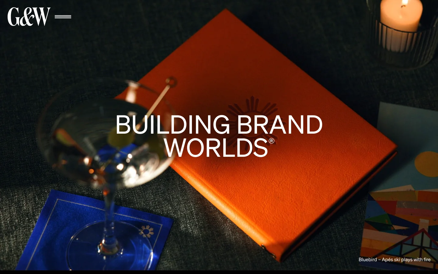 The desktop homepage features a dark, sophisticated aesthetic with a large hero image of a cocktail and an orange book titled 'Building Brand Worlds®'. A navigation menu is at the top left, and a grid of high-quality project photography follows, showcasing various branding and packaging works.