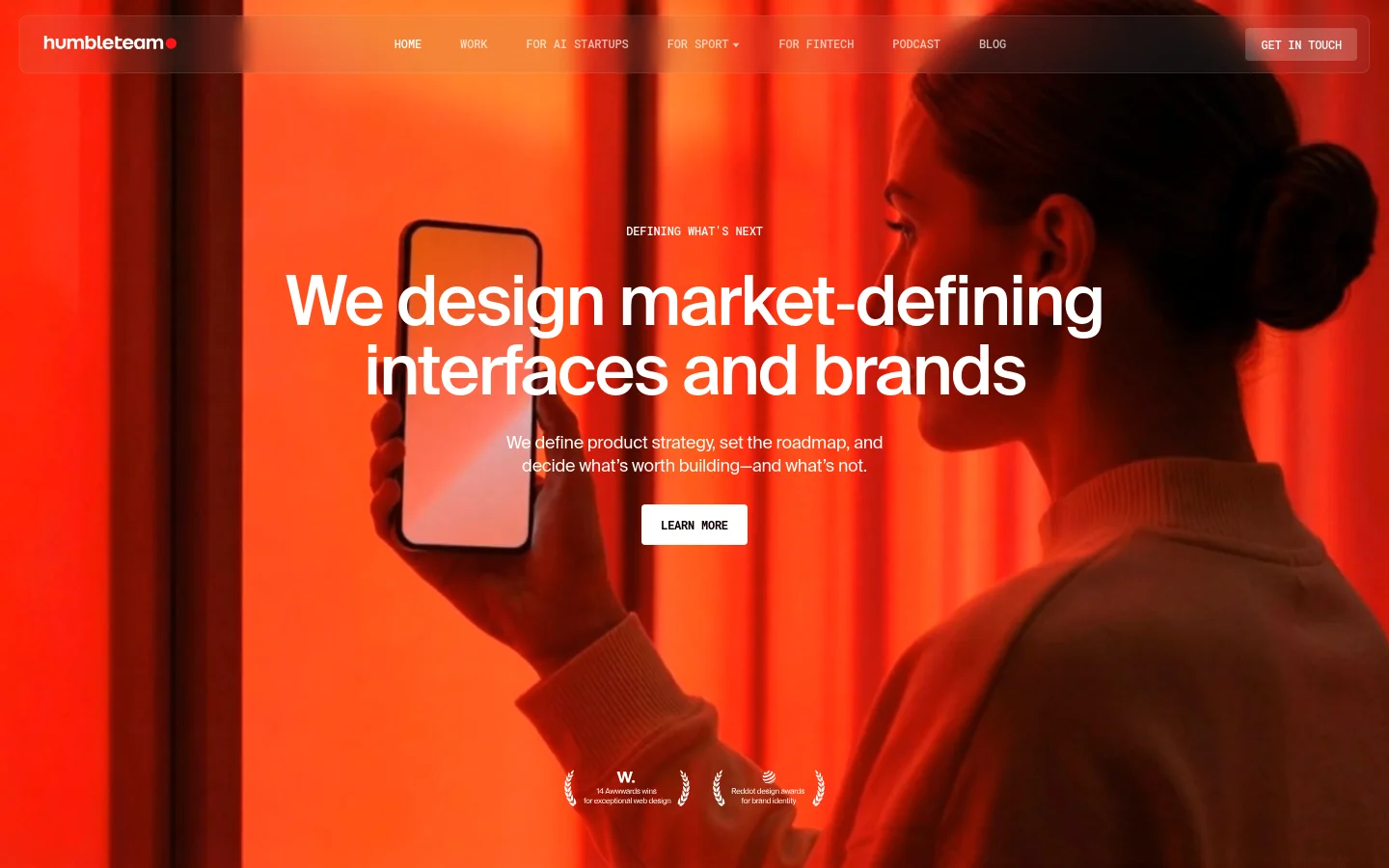 The desktop homepage features a large hero video of a person holding a smartphone against a vibrant red background. A navigation bar at the top includes links for Work, AI Startups, Sport, and Fintech, with a 'Get in Touch' button on the right. The central headline reads 'We design market-defining interfaces and brands' above a 'Learn More' button.
