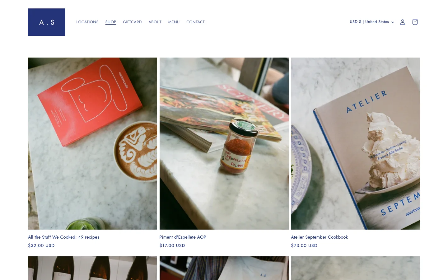 The desktop shop page features a clean, minimalist layout with a white background and a dark blue header containing navigation links like 'LOCATIONS' and 'SHOP'. A grid of large, high-quality lifestyle product images displays items such as cookbooks and spices, each accompanied by a title and price in black text. The design uses ample white space to create an elegant, editorial aesthetic.