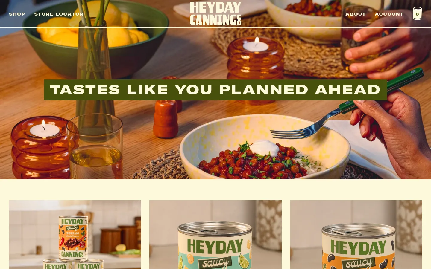 The desktop page features a large hero image of a warm, set dinner table with the headline 'TASTES LIKE YOU PLANNED AHEAD' in white text on a green banner. Below, a grid of product collections displays colorful canned goods and prepared meals, including a Saucy Bean Variety Pack and various 6-packs. The navigation bar is at the top with links for Shop, Store Locator, About, and Account.