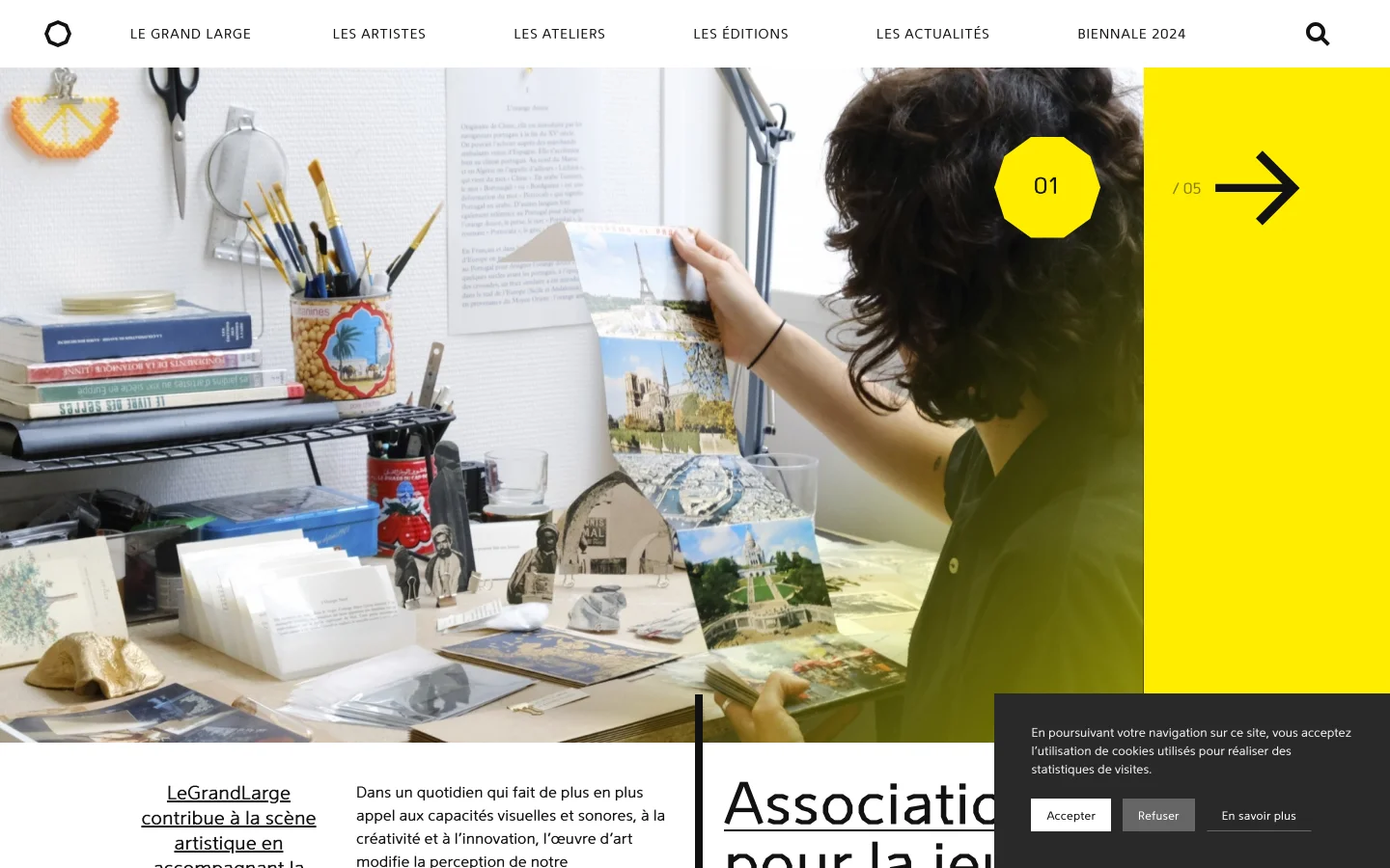 The desktop homepage features a clean white and yellow color scheme with a top navigation menu and a large hero image slider. The current slide shows a person's hands working with photographs on a desk filled with art supplies, overlaid with a large yellow geometric shape containing slide numbers and a navigation arrow. Below the image, large black serif text introduces the association, accompanied by descriptive paragraphs.