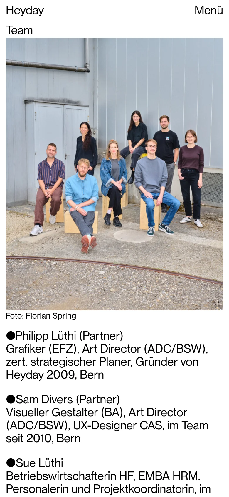 The mobile version presents a vertical layout with the team photo spanning the full width of the screen. Below the image, the team member names and detailed professional biographies are listed in a single column. The navigation is condensed into a 'Menü' button at the top right.