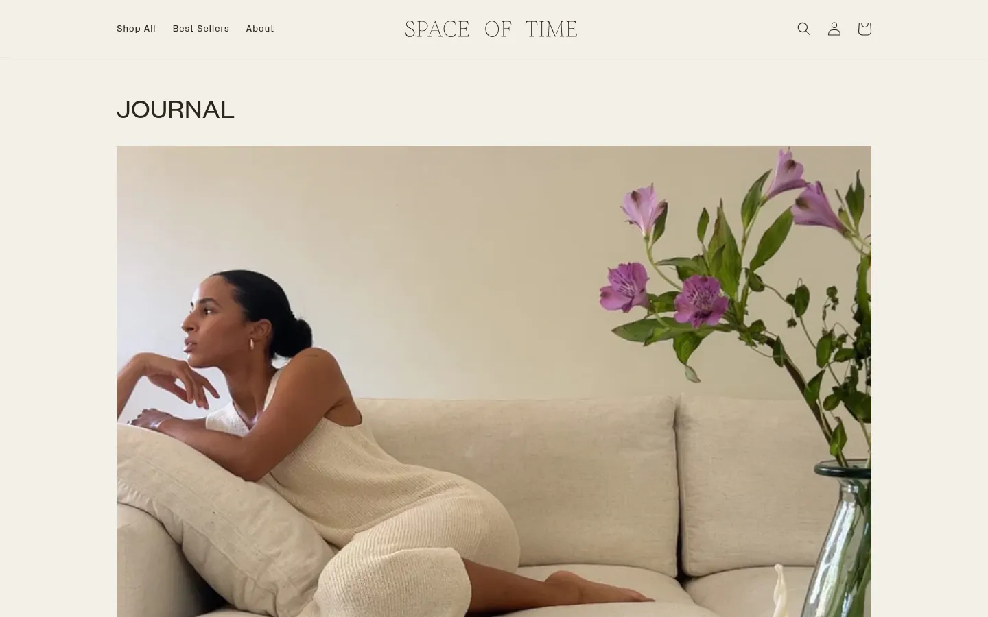 The Journal page features a minimalist design with a large hero image of a woman reclining on a neutral-toned sofa next to purple flowers. The layout includes a clean white header with navigation links for 'Shop All', 'Best Sellers', and 'About', alongside search, account, and cart icons. Below the hero section, blog posts are arranged in a vertical feed with large images, bold headlines, and brief text excerpts.