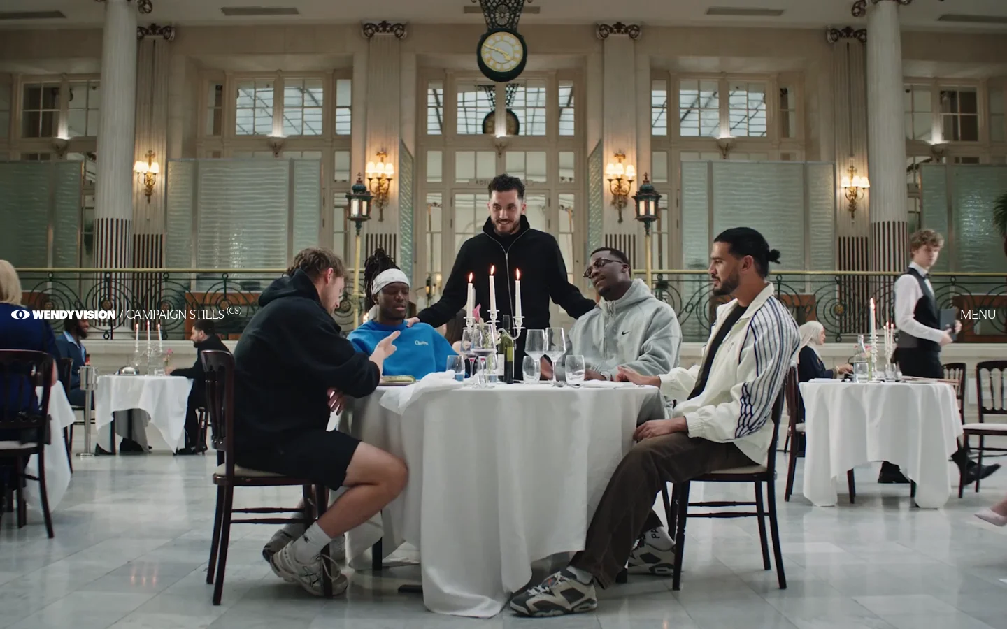 The desktop homepage features a large cinematic hero image of a group of diverse people seated around a white-clothed dinner table in a grand, high-ceilinged hall. The WENDYVISION logo is in the top left, with navigation links for 'Service Production', 'Campaign Stills', and 'Menu' spread across the top and side. The color palette is warm and sophisticated with neutral tones.