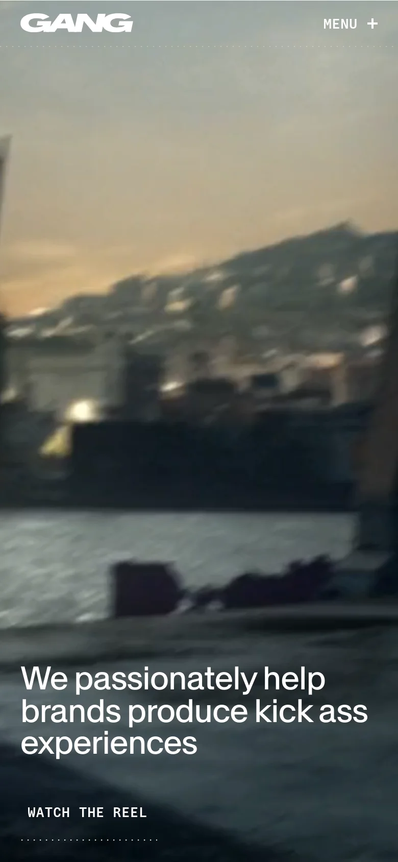 The mobile version maintains the dark aesthetic but optimizes the layout for a vertical screen, showing a tall, blurred landscape hero image. The main headline and 'Watch the reel' button are centered and stacked vertically. The navigation is simplified with a 'MENU +' icon in the top right, and the content sections follow a vertical scrolling format.