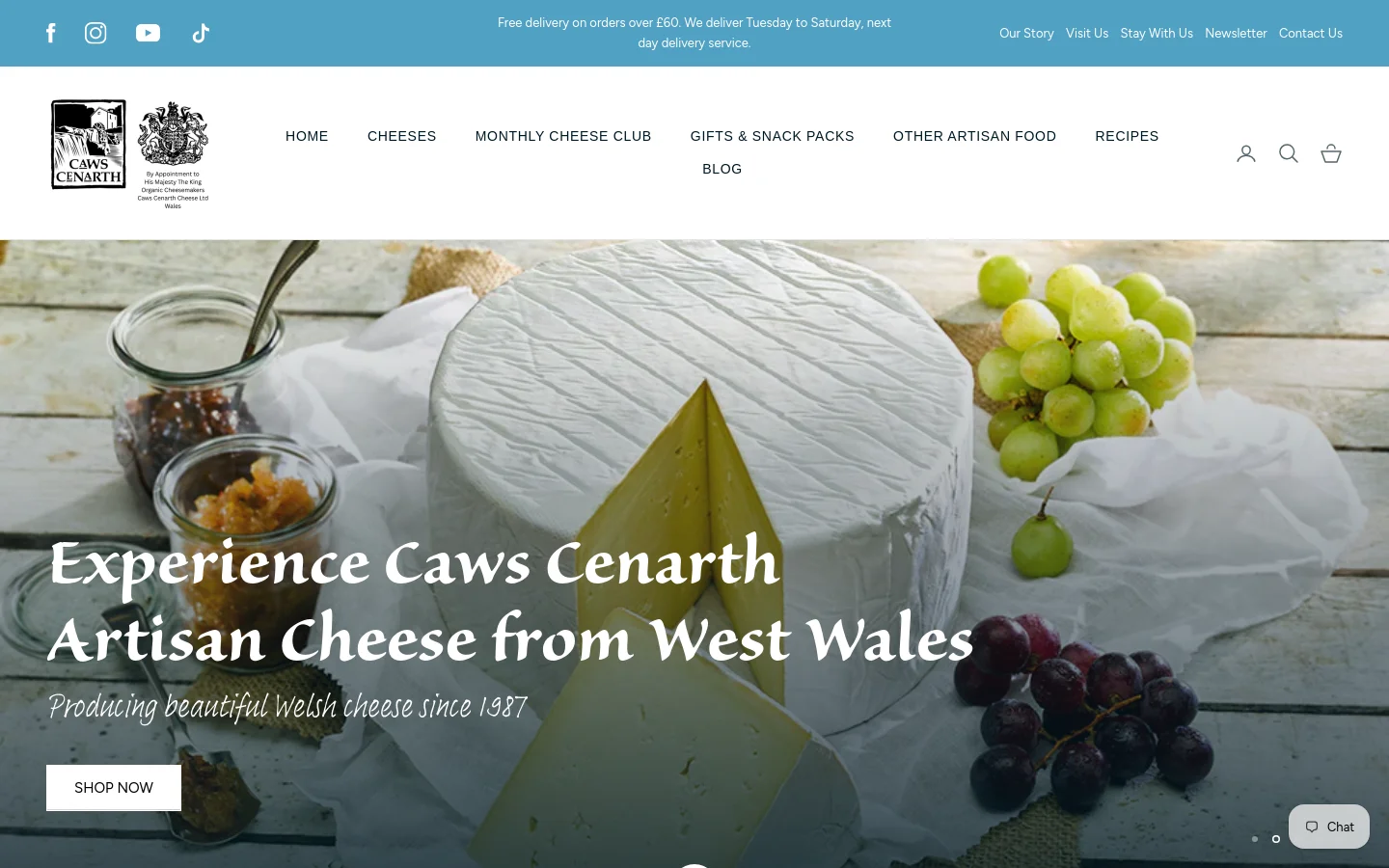 The desktop homepage features a large hero image of a cheese wheel with grapes and preserves, overlaid with the headline 'Experience Caws Cenarth Artisan Cheese from West Wales' and a 'SHOP NOW' button. A blue announcement bar at the top provides delivery information, and a white navigation menu includes links for Home, Cheeses, and Monthly Cheese Club. The layout is spacious with a clean white background for the header and navigation.