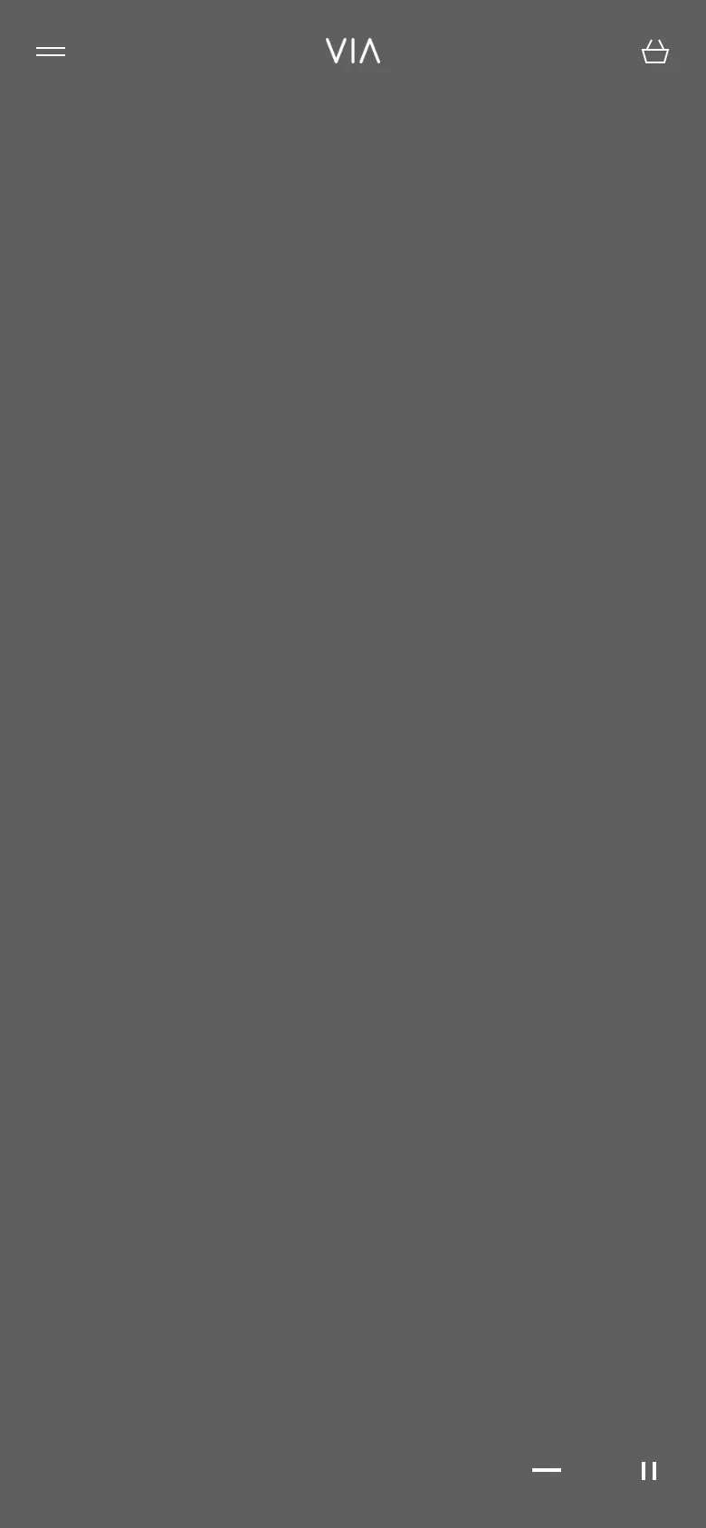 The mobile version maintains the minimalist dark gray aesthetic and centered VIA logo but simplifies the header to a hamburger menu icon on the left and a shopping bag icon on the right. The layout is vertically oriented, focusing on the large hero image area with playback controls at the bottom.
