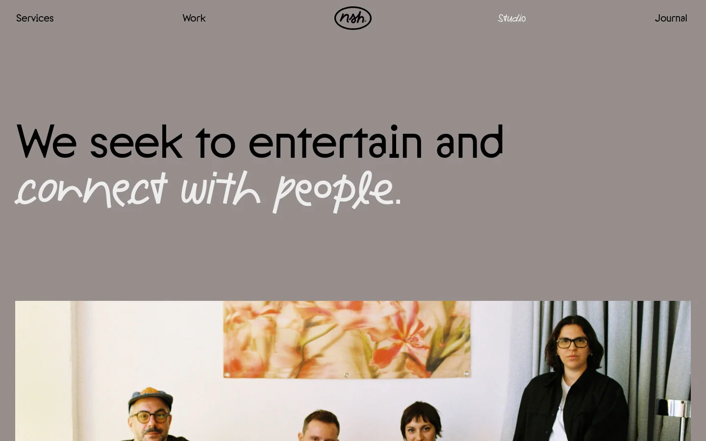 The desktop page features a minimalist design with a gray background and large, bold typography. A hero section displays the headline 'We seek to entertain and connect with people' in black and white stylized fonts, positioned above a wide photograph of a creative team sitting on a sofa in a bright studio. A navigation bar at the top includes links for Services, Work, Studio, and Journal, flanking a central circular logo.