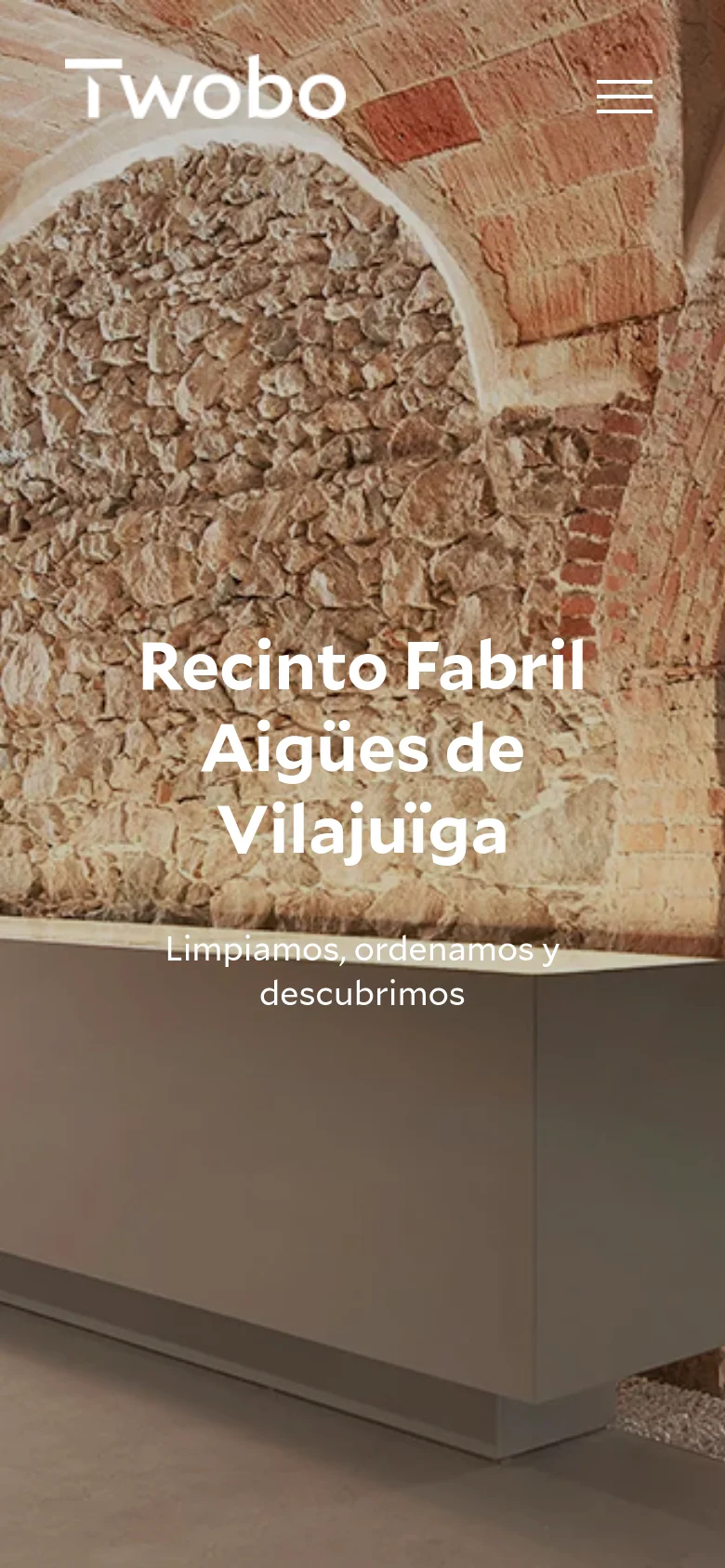 The mobile version uses a full-screen hero image of a stone archway with the headline 'Recinto Fabril Aigües de Vilajuïga' centered in white text. The navigation is condensed into a hamburger menu icon in the top right, and the 'Twobo' logo is positioned in the top left against the textured background.