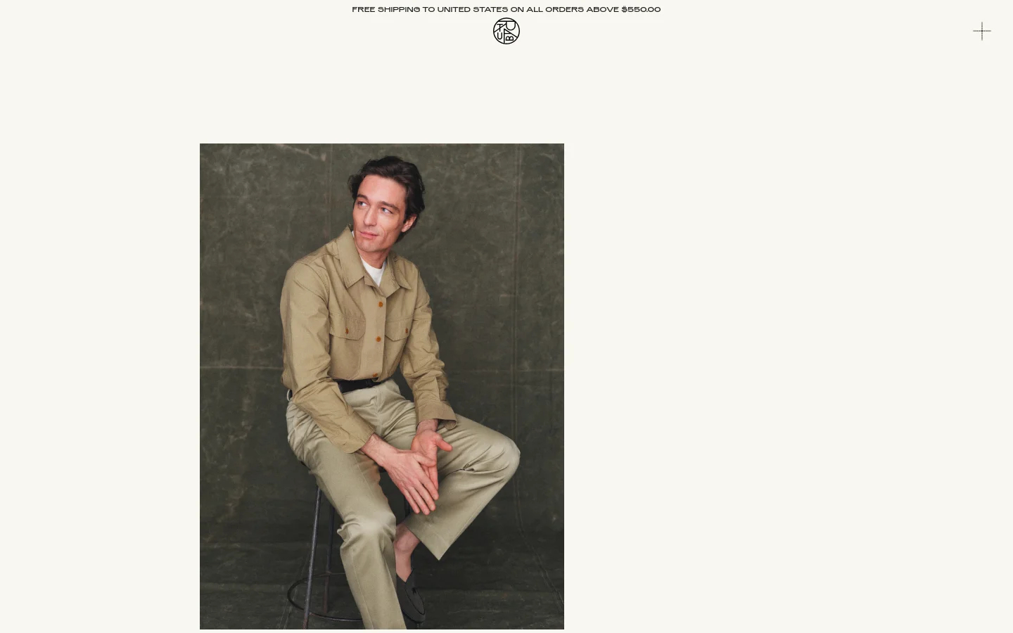 The desktop homepage features a minimalist design with a cream background and a large hero image of a man sitting on a stool wearing tan clothing. A thin announcement bar at the top reads 'FREE SHIPPING TO UNITED STATES ON ALL ORDERS ABOVE $550.00', flanked by a central logo and a plus icon. Below the hero image, sections for 'LATEST', 'EXPLORE OUR JOURNAL', and 'CORE COLLECTION' are arranged vertically with large headings and high-quality photography.