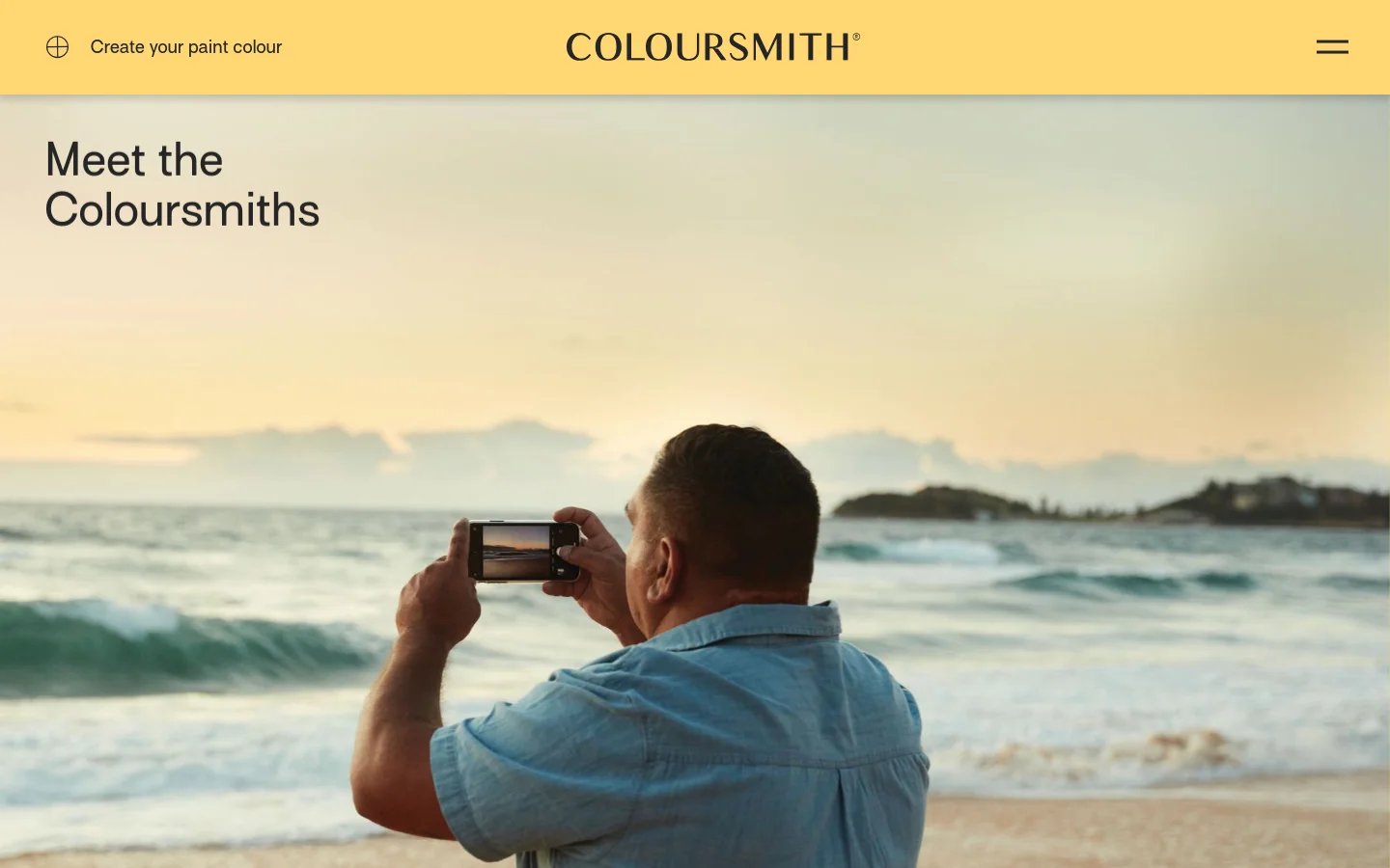 The desktop page features a yellow header with the Coloursmith logo and a 'Create your paint colour' link. A large hero image shows a man from behind taking a photo of a sunset beach scene, overlaid with the headline 'Meet the Coloursmiths'. Below the hero, a grid layout displays individual color stories, each featuring a color swatch, a title, an author, and a short testimonial.