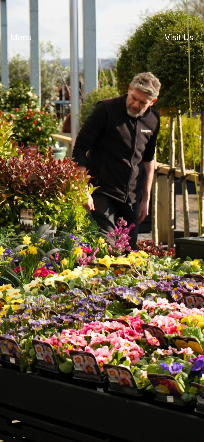 The mobile version displays the same hero image of colorful flowers and a gardener, but the layout is vertically oriented. The 'Menu' and 'Visit Us' text links are placed prominently at the top of the screen to accommodate touch navigation.
