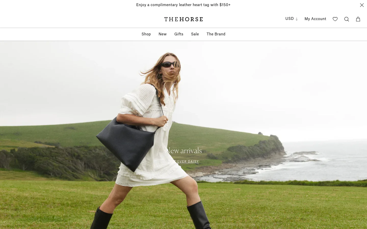 The desktop homepage for The Horse features a large hero image of a woman in a white dress and black boots carrying a black leather bag against a coastal landscape. A navigation menu sits at the top with links for Shop, New, Gifts, Sale, and The Brand, alongside a promotional banner for a free leather heart tag. Below the hero, a grid of product images showcases collections like Tilly, Fin, Large Daisy, and Clementine.