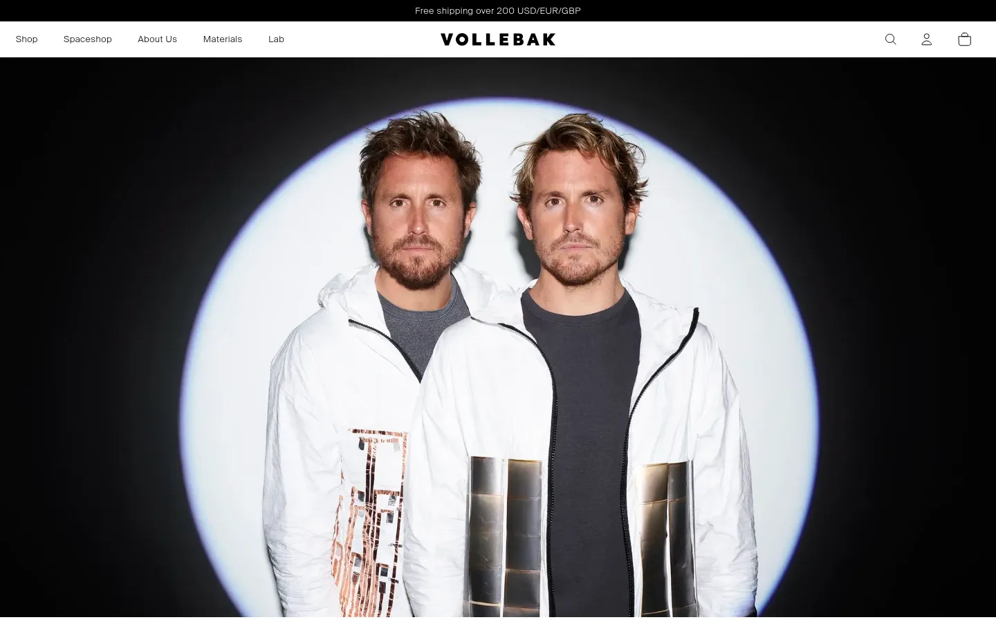 The desktop About Us page features a minimalist white header with navigation links for Shop, Spaceshop, About Us, Materials, and Lab, alongside search, account, and bag icons. A large hero image shows two men, the founders, wearing white technical jackets against a circular spotlight background. The layout is clean with high-contrast black and white tones.