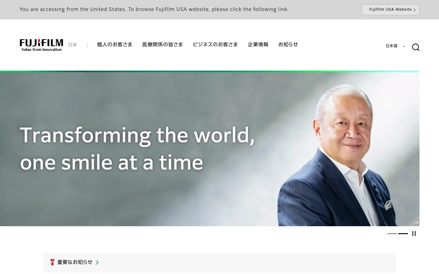 The Fujifilm Japan homepage features a large hero banner with a smiling man and the headline 'Transforming the world, one smile at a time' against a light blue background. Below the banner, a white notification bar highlights important news, followed by a grid of large image tiles for Consumer, Healthcare, and Business segments. The layout is clean with a white header containing the Fujifilm logo, navigation links, and a search icon.