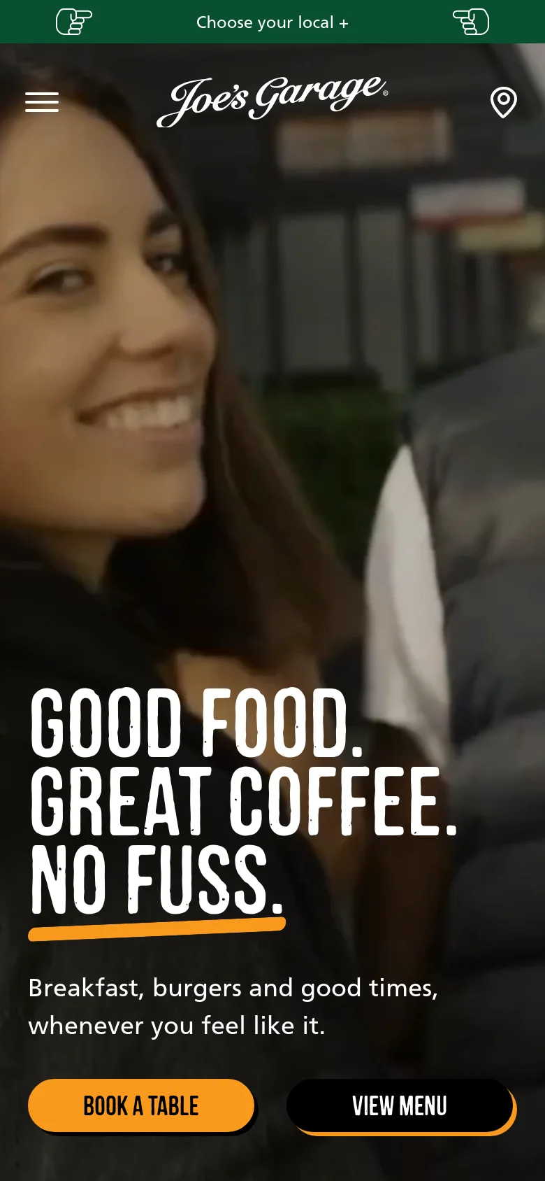 The mobile version uses a vertically oriented hero image of a smiling woman, with the same bold white headline and subtext centered on the screen. The navigation is condensed into a hamburger menu and a location icon, while the primary 'Book a Table' and 'View Menu' calls to action are prominent orange and black buttons at the bottom.