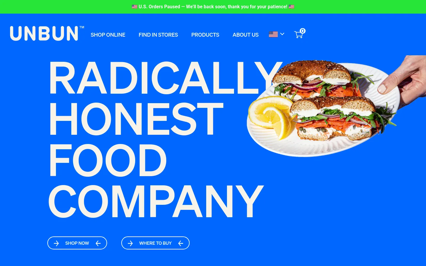 The desktop homepage features a bright blue background with large, bold white typography. The hero section displays the headline 'RADICALLY HONEST FOOD COMPANY' alongside a high-quality image of a bagel sandwich with salmon and lemon on a white plate. Two pill-shaped buttons, 'SHOP NOW' and 'WHERE TO BUY', are positioned at the bottom of the hero section under a navigation menu.