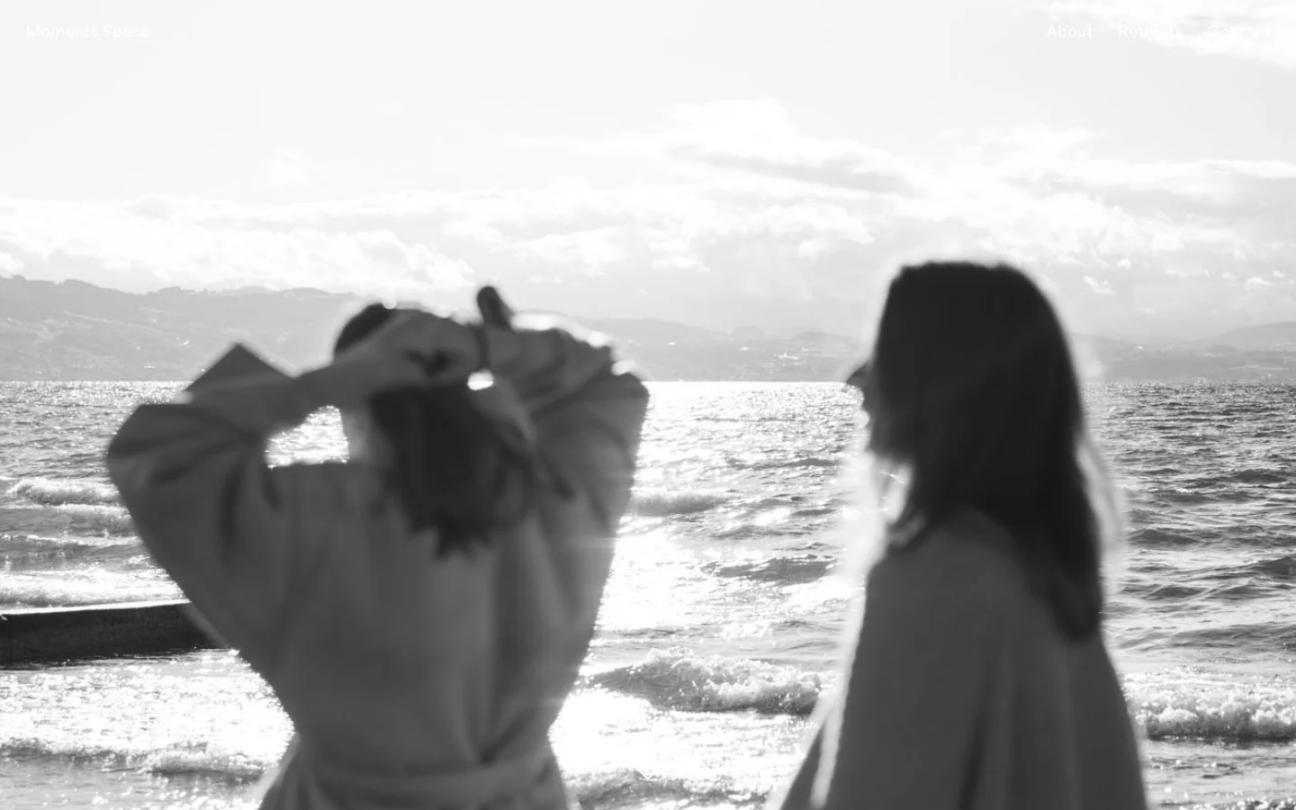 The desktop About page features a large, black-and-white hero image of two women looking out over a sparkling sea toward distant mountains. A minimalist navigation menu with links for About, Retreats, and Contact is positioned at the top, overlaid on the bright sky. The layout uses clean typography with large headings and ample white space for a calm, reflective feel.