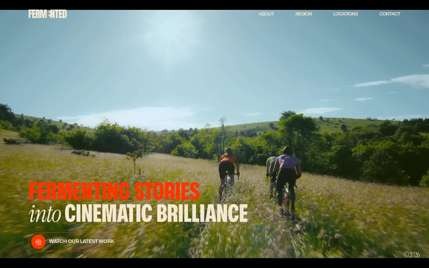 The desktop homepage features a full-width cinematic hero video of cyclists in a sunny meadow. The headline 'FERMENTING STORIES into CINEMATIC BRILLIANCE' is overlaid in bold red and white text, with a prominent 'WATCH OUR LATEST WORK' button below. A white navigation bar at the top includes the Fermented logo and links for About, Region, Locations, and Contact.