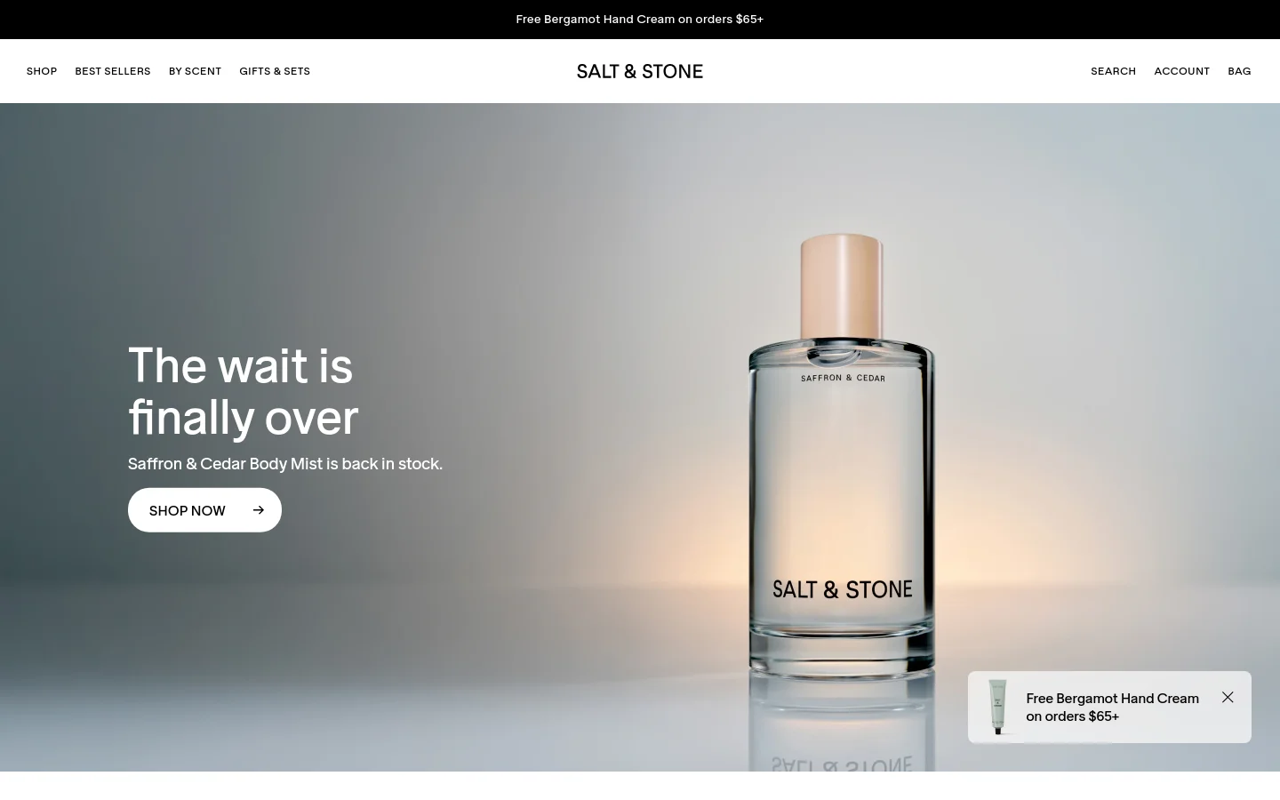 The desktop homepage features a minimalist design with a large hero image of a Saffron & Cedar Body Mist bottle against a soft, gradient background. A prominent headline reads 'The wait is finally over' with a 'SHOP NOW' call-to-action button. A thin announcement bar at the top offers free Bergamot Hand Cream on orders over $65.