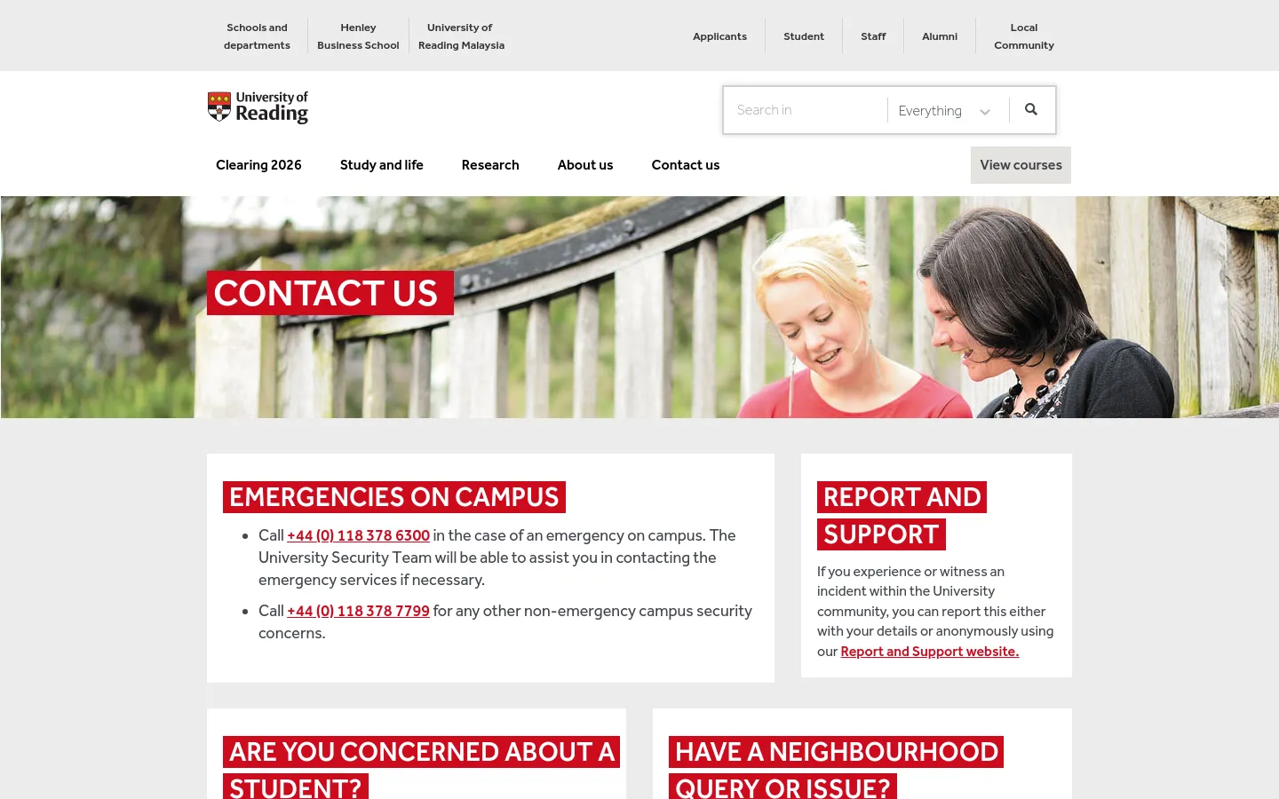 The University of Reading Contact Us page features a wide hero image of two smiling women outdoors, overlaid with a red 'CONTACT US' banner. Below, the layout uses a white background with large red headings for 'EMERGENCIES ON CAMPUS' and 'REPORT AND SUPPORT', organized into a clean grid. The header includes the university logo, a search bar, and a navigation menu with links like 'Clearing 2026' and 'Research'.