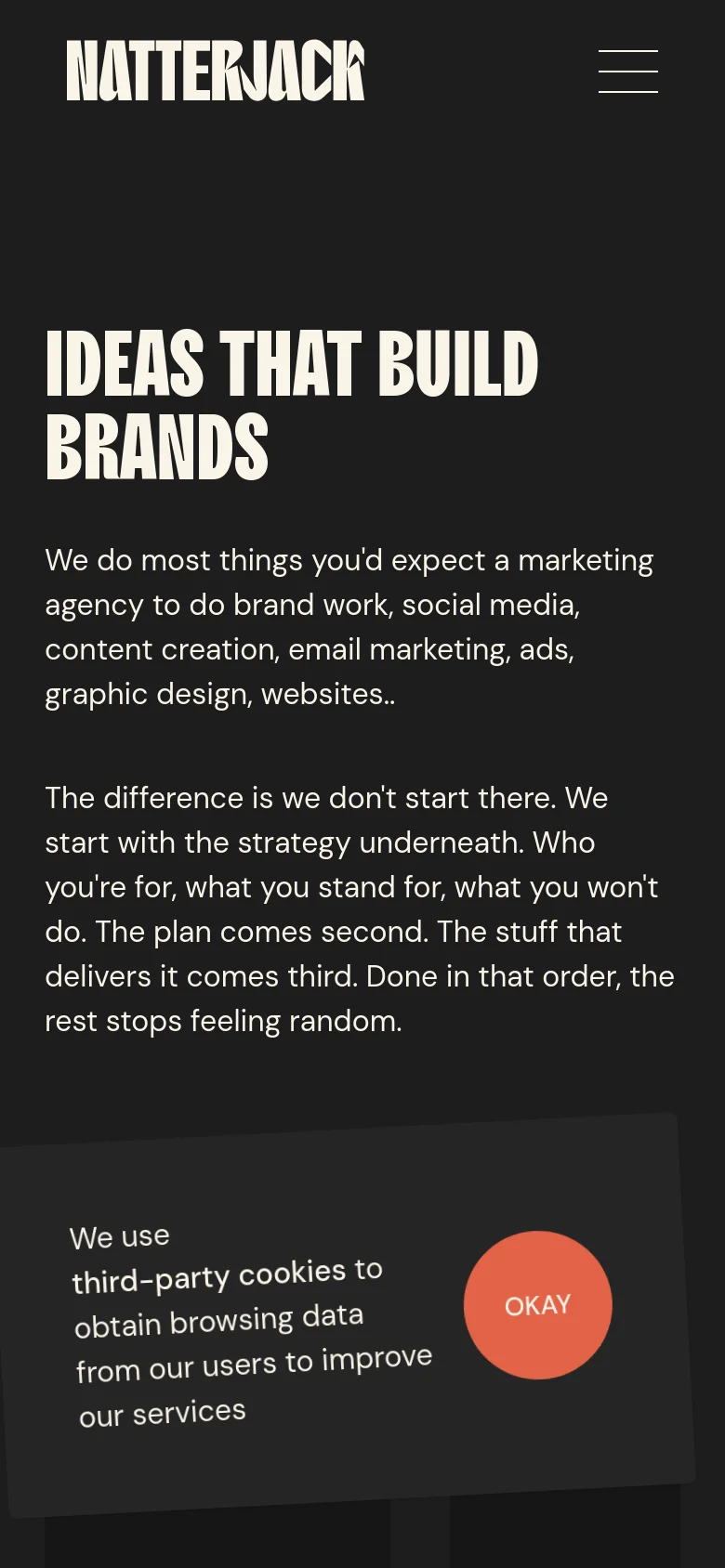 Mobile version of the 'What We Do' page with a simplified vertical layout on a dark background. The large white headline 'IDEAS THAT BUILD BRANDS' is followed by several paragraphs of text, and the navigation is collapsed into a hamburger menu icon at the top right. A large circular orange 'OKAY' button for cookie consent is prominent at the bottom.