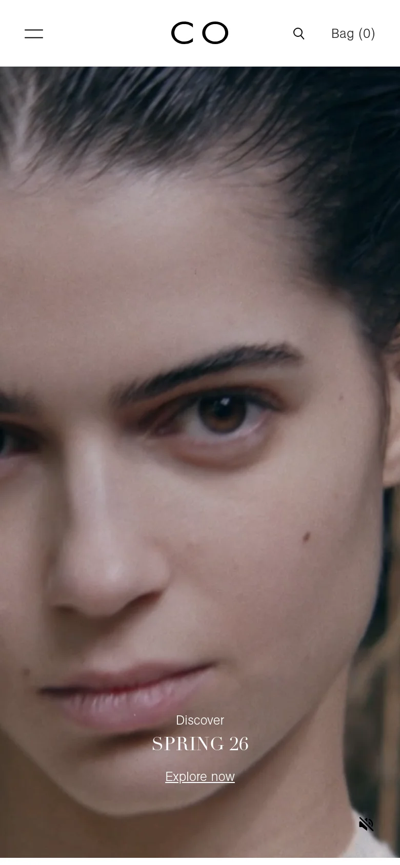 The mobile version presents a streamlined vertical layout with a centered logo and a hamburger menu icon on the left. The hero image remains a prominent close-up of a woman's face, with the 'Discover SPRING 26' text and 'Explore now' link positioned centrally near the bottom of the frame. The navigation is condensed, prioritizing the search icon and shopping bag status at the top.