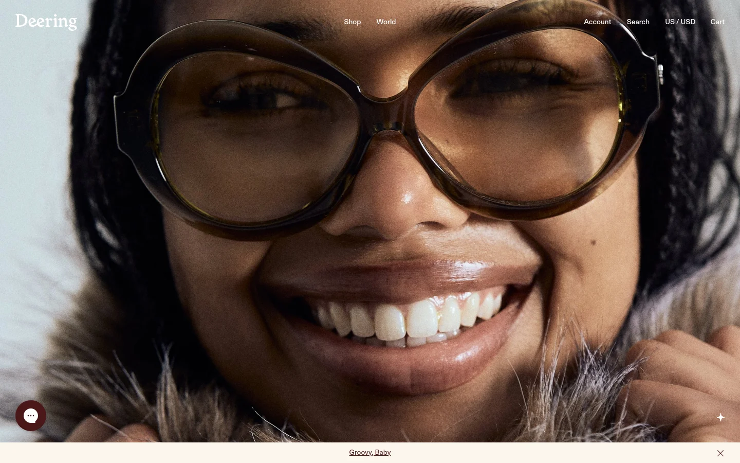 The desktop homepage features a large, close-up hero image of a smiling person wearing oversized tortoiseshell glasses and a faux fur collar. The brand name 'Deering' is in the top left, with navigation links for Shop, World, Account, Search, Currency, and Cart spread across the top. A secondary section below features the headline 'You hold the universe in your hands' with buttons for 'Groovy, Baby' and 'New Arrivals'.