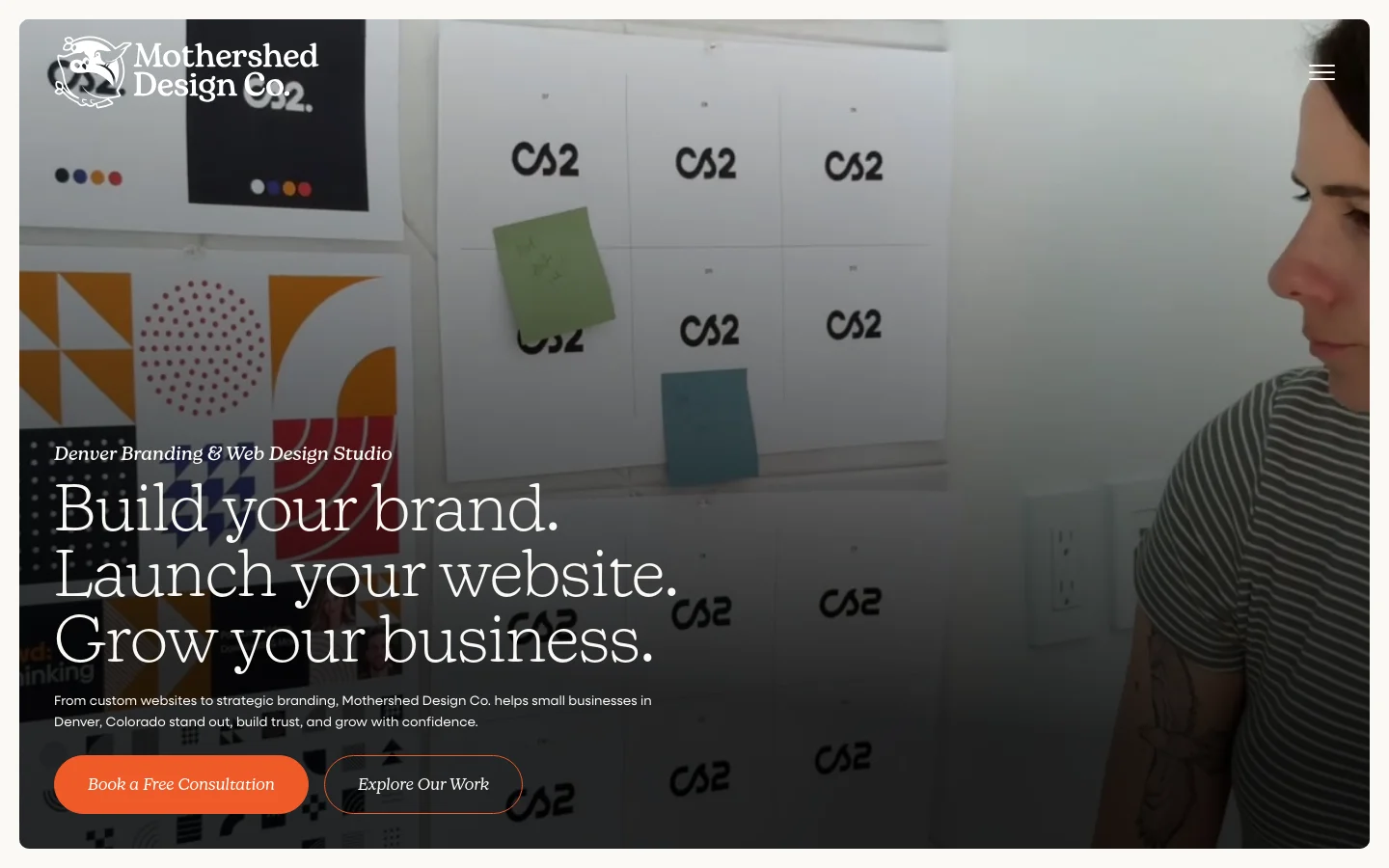 The desktop homepage features a large hero image of a designer working in a studio, overlaid with white serif text that reads 'Build your brand. Launch your website. Grow your business.' A primary orange call-to-action button says 'Book a Free Consultation' next to an outlined 'Explore Our Work' button. The layout is clean with a dark overlay on the image to ensure text readability.