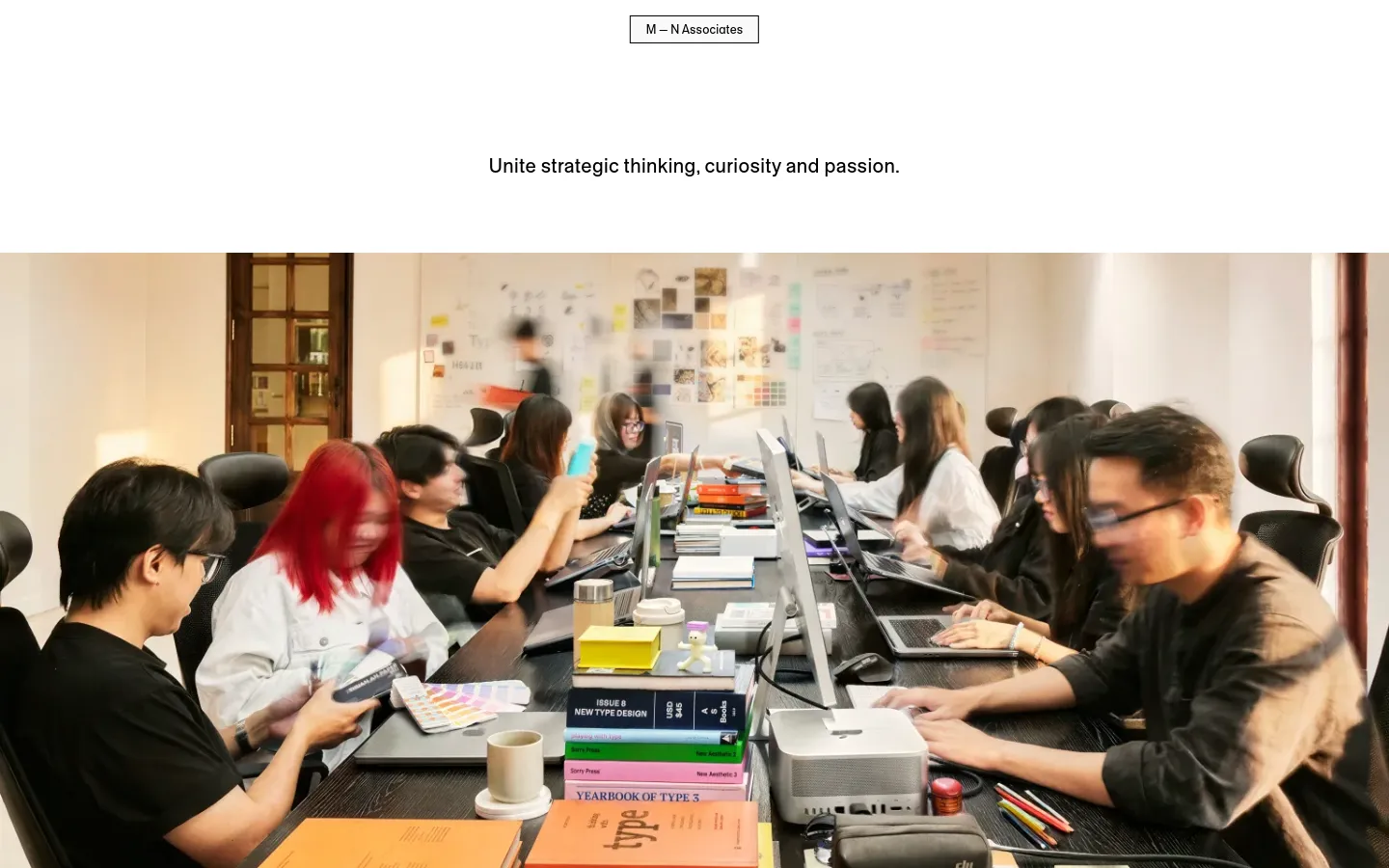 The desktop page features a clean, minimalist white layout with centered black text. A large hero image shows a creative team working around a long table filled with design books, laptops, and color palettes in a bright studio. The header contains a simple 'M — N Associates' logo button.