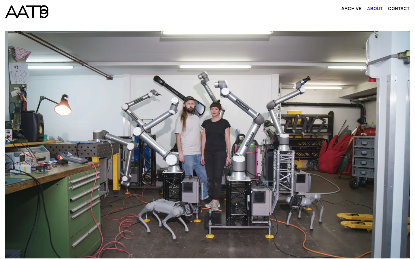 The desktop page features a minimalist white layout with the AATB logo on the top left and navigation links on the top right. A large hero image shows two artists standing in a workshop surrounded by robotic arms and technical equipment. Below the image, several paragraphs of black text on a white background describe the duo's practice, exhibitions, and collaborators.