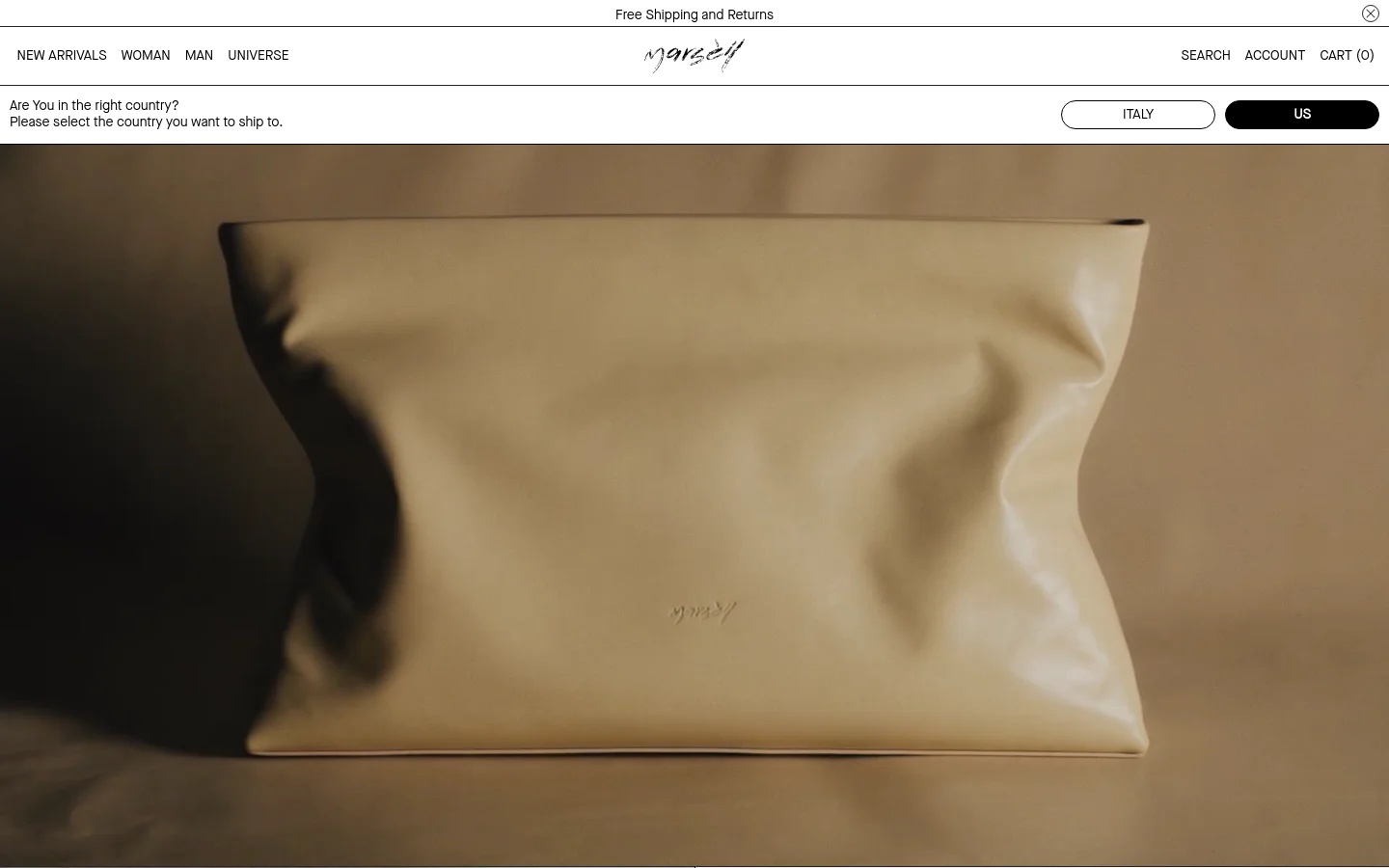 The desktop homepage features a minimalist design with a large, high-resolution hero image of a cream-colored leather clutch against a neutral background. A top navigation bar includes links for New Arrivals, Woman, Man, and Universe, alongside search, account, and cart options. A banner at the top offers free shipping and returns and includes a country selection prompt.