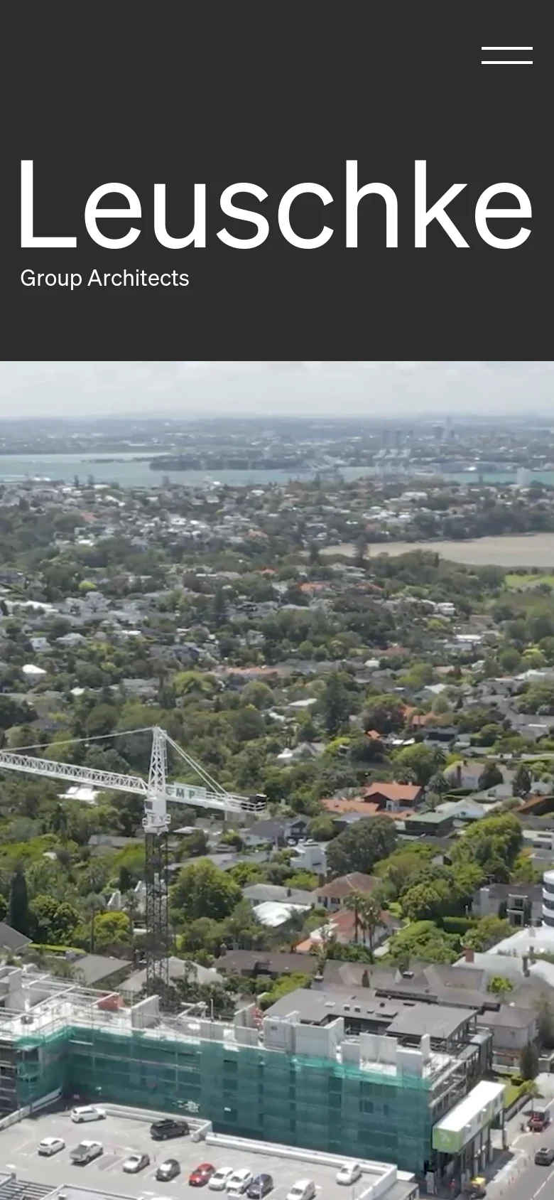 The mobile version simplifies the hero section into a vertical stack, placing the large white 'Leuschke Group Architects' title over a full-width aerial photograph of a coastal cityscape. The dark grey background is reduced to a header area, and the navigation is condensed into a hamburger menu icon at the top right.